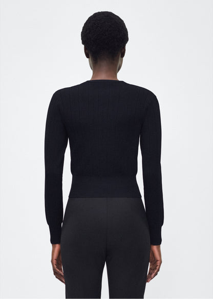 Image of model facing back P25625UB, Faye Cardigan in black, Adam Lippes P25 collection: A classic pointelle stitch cardigan, crafted in our signature compact jacquard. Designed with a fitted bodice and delicate stitching, the piece is finished with covered buttons along the front placket for a refined look.