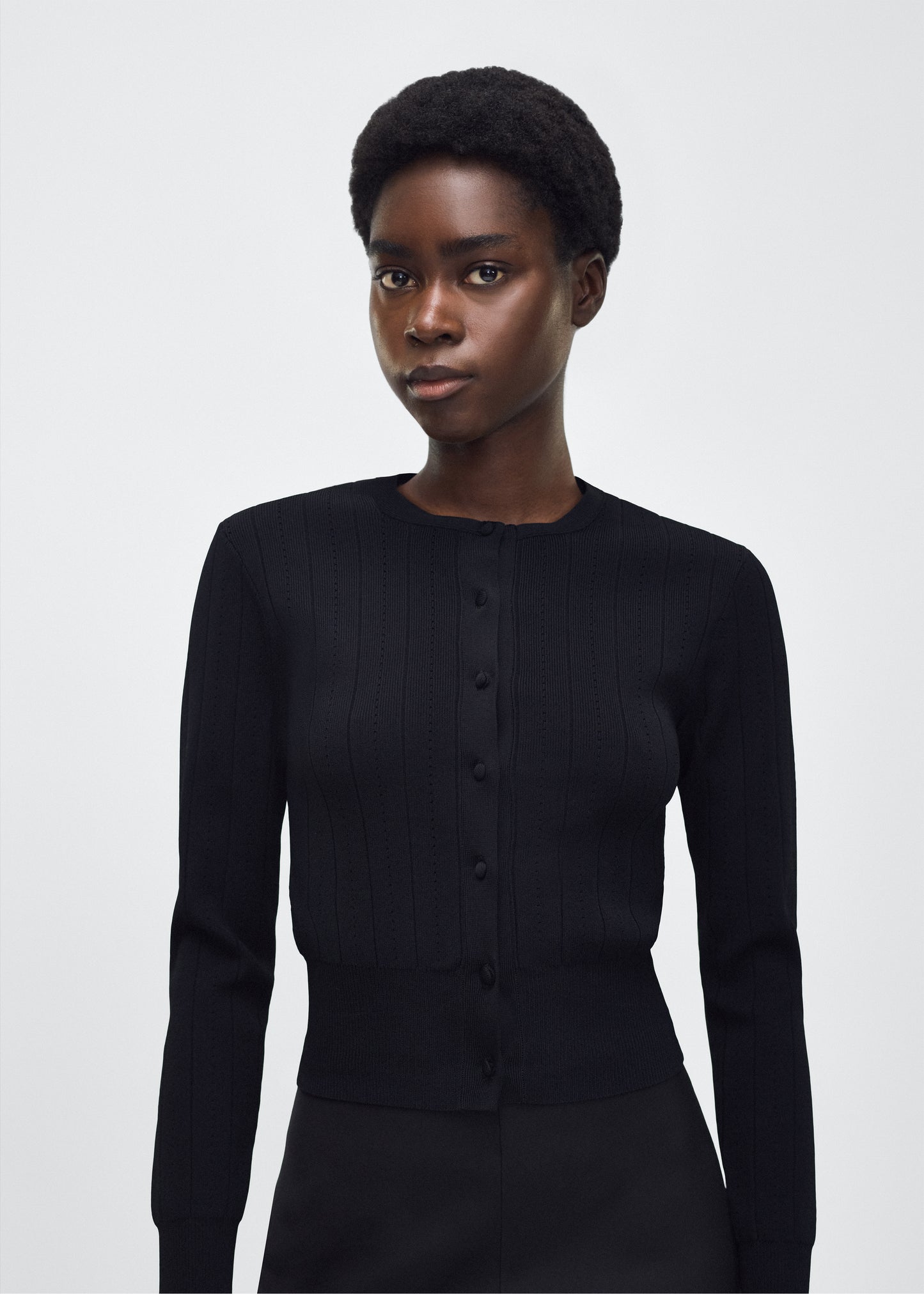Image of model facing front, P25625UB, Faye Cardigan in black, Adam Lippes P25 collection: A classic pointelle stitch cardigan, crafted in our signature compact jacquard. Designed with a fitted bodice and delicate stitching, the piece is finished with covered buttons along the front placket for a refined look.