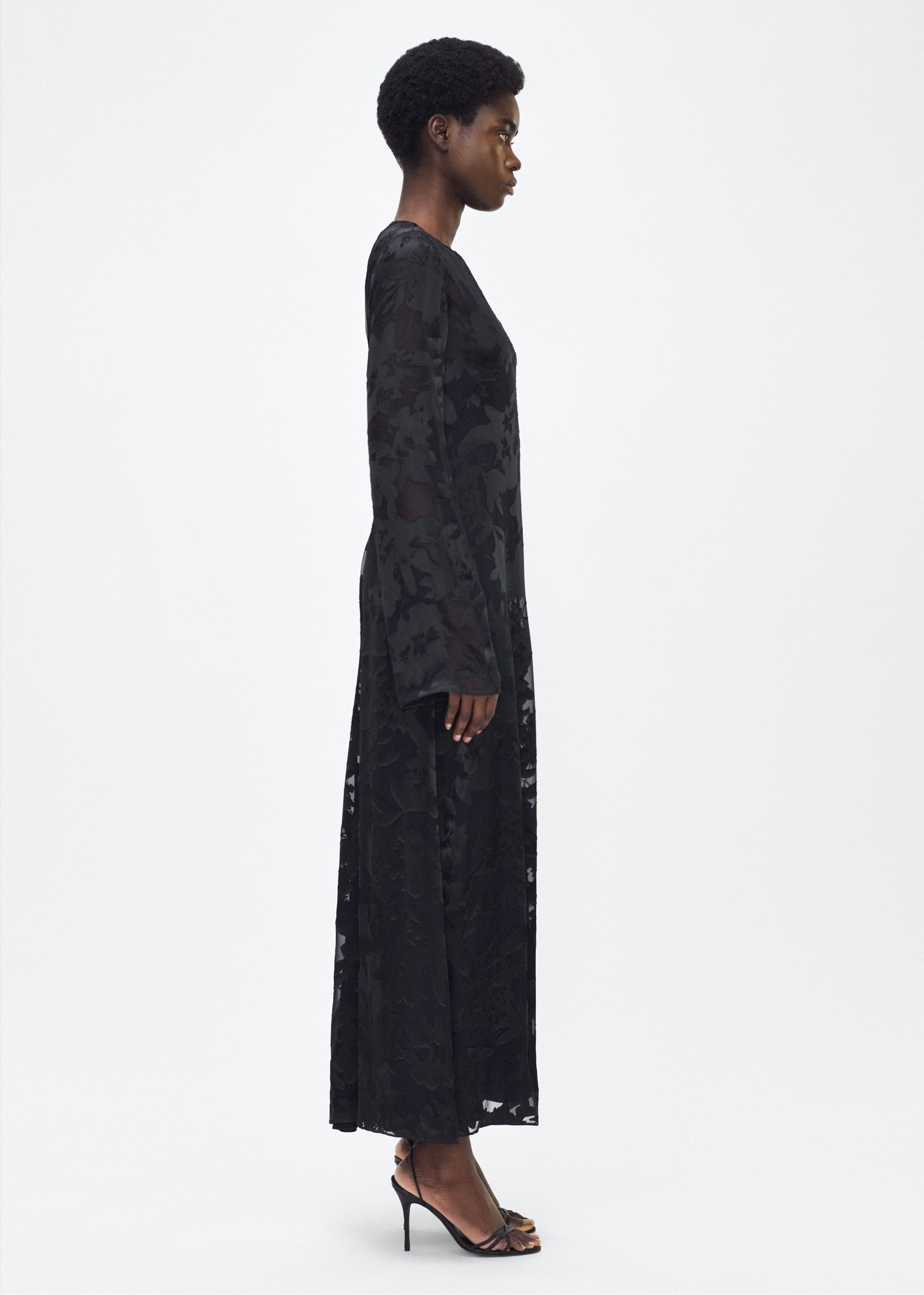 Model facing side - The Sylvia Dress in Black, Adam Lippes P25 Collection: The Sylvia Dress in black Devore features a dramatic back cut-out, adding an alluring touch to the dress’s richly textured floral design. Complete with a detachable silk cami dress, this stunning piece offers both versatility and timeless sophistication.