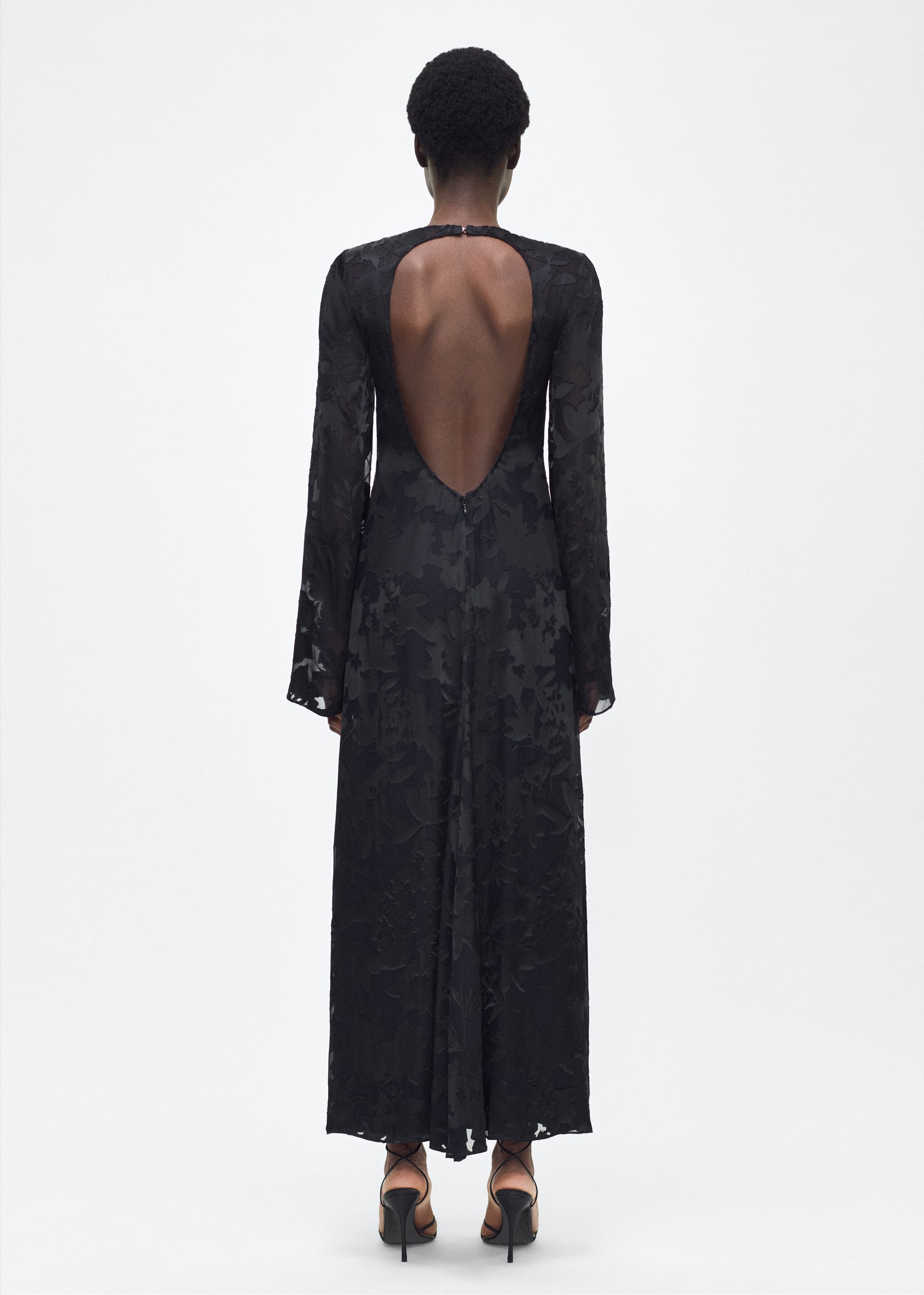 Model facing back - The Sylvia Dress in Black, Adam Lippes P25 Collection: The Sylvia Dress in black Devore features a dramatic back cut-out, adding an alluring touch to the dress’s richly textured floral design. Complete with a detachable silk cami dress, this stunning piece offers both versatility and timeless sophistication.