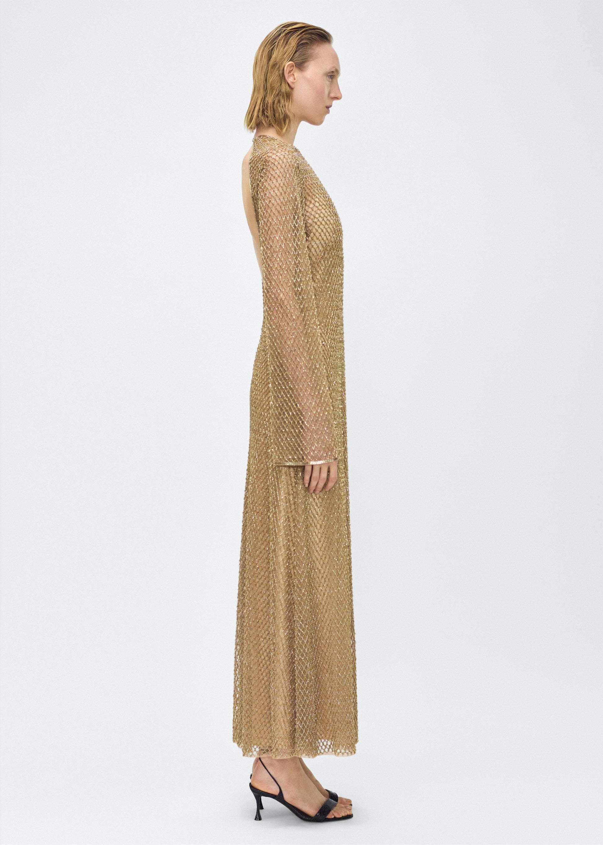 Model facing side Image of the Sylvia Dress in Gold, P25 Adam Lippes collection; The Sylvia Dress in gold glass beaded tulle features intricate hand-beading, a process that takes multiple days, arranged in the signature diamond motif pattern for a striking visual effect. Designed with an elegant open back and a detachable cami, this dress offers versatile styling options with maximum impact.