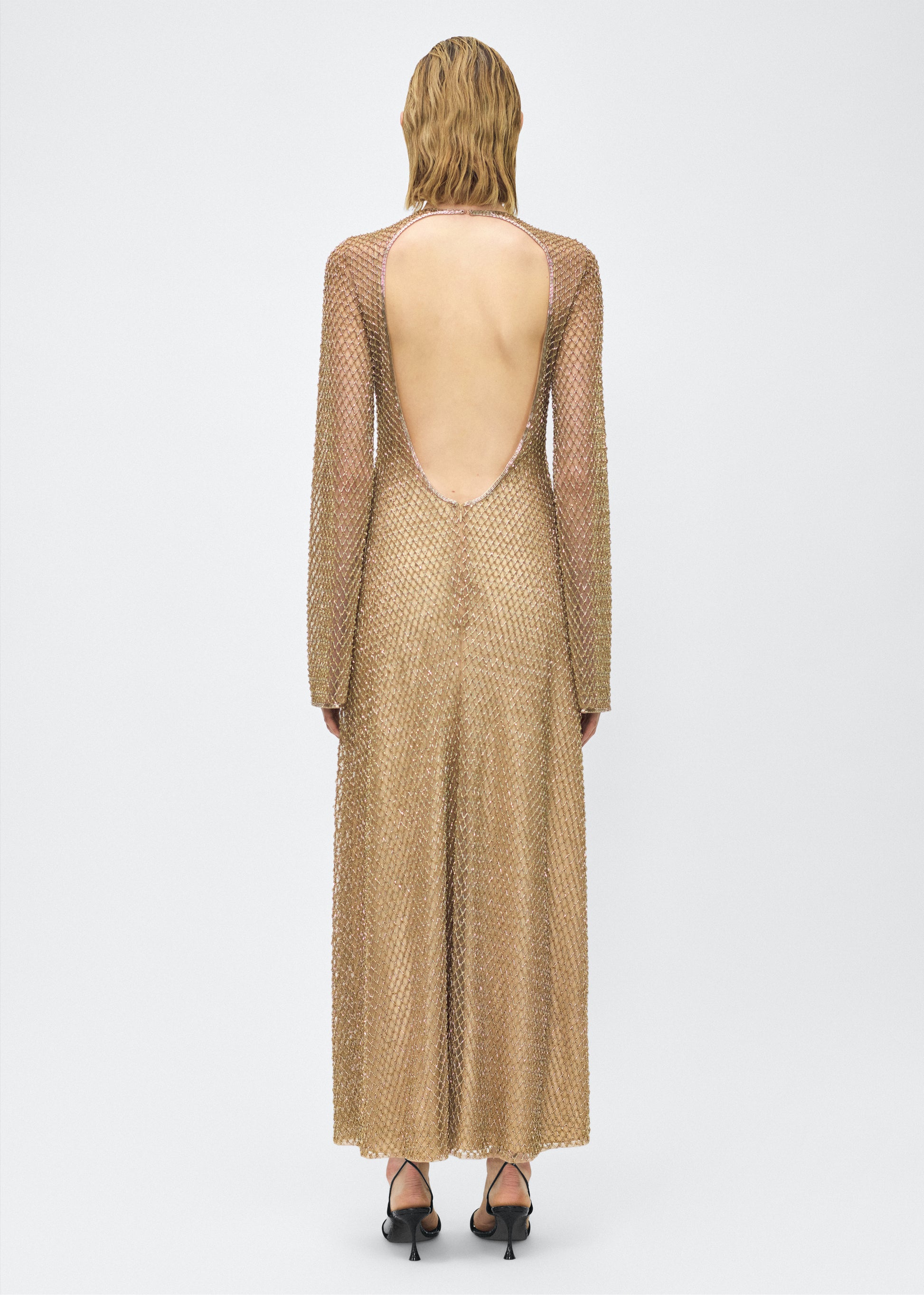 Model facing back, Image of the Sylvia Dress in Gold, P25 Adam Lippes collection; The Sylvia Dress in gold glass beaded tulle features intricate hand-beading, a process that takes multiple days, arranged in the signature diamond motif pattern for a striking visual effect. Designed with an elegant open back and a detachable cami, this dress offers versatile styling options with maximum impact.
