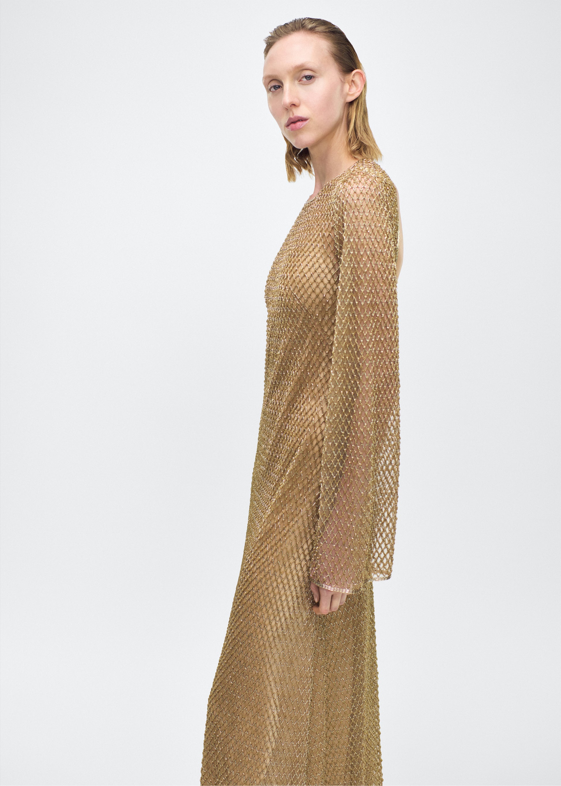 Model facing side Image of the Sylvia Dress in Gold, P25 Adam Lippes collection; The Sylvia Dress in gold glass beaded tulle features intricate hand-beading, a process that takes multiple days, arranged in the signature diamond motif pattern for a striking visual effect. Designed with an elegant open back and a detachable cami, this dress offers versatile styling options with maximum impact.