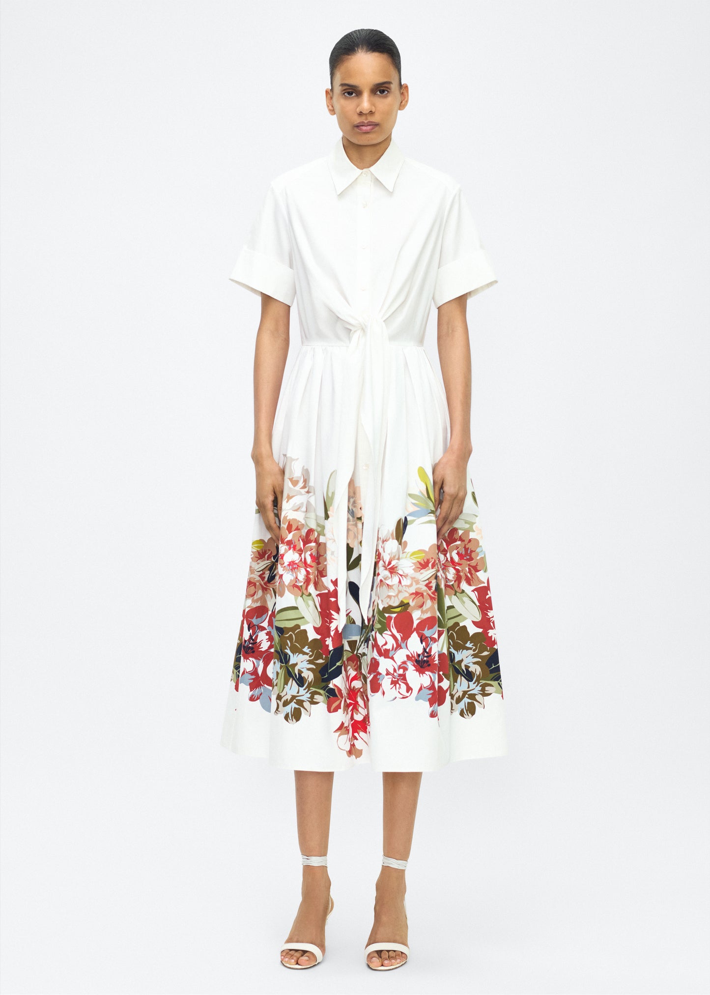 Model facing front, Attacher Dress in white multi, P25 Collection from Adam Lippes; Our classic Attacher silhouette returns, crafted from Italian cotton poplin and printed with this season’s Sicily-inspired floral motif. Featuring side pockets and diamond monogram mother of pearl buttons, the attached waist sash can be tied at the front or back to cinch the waist.