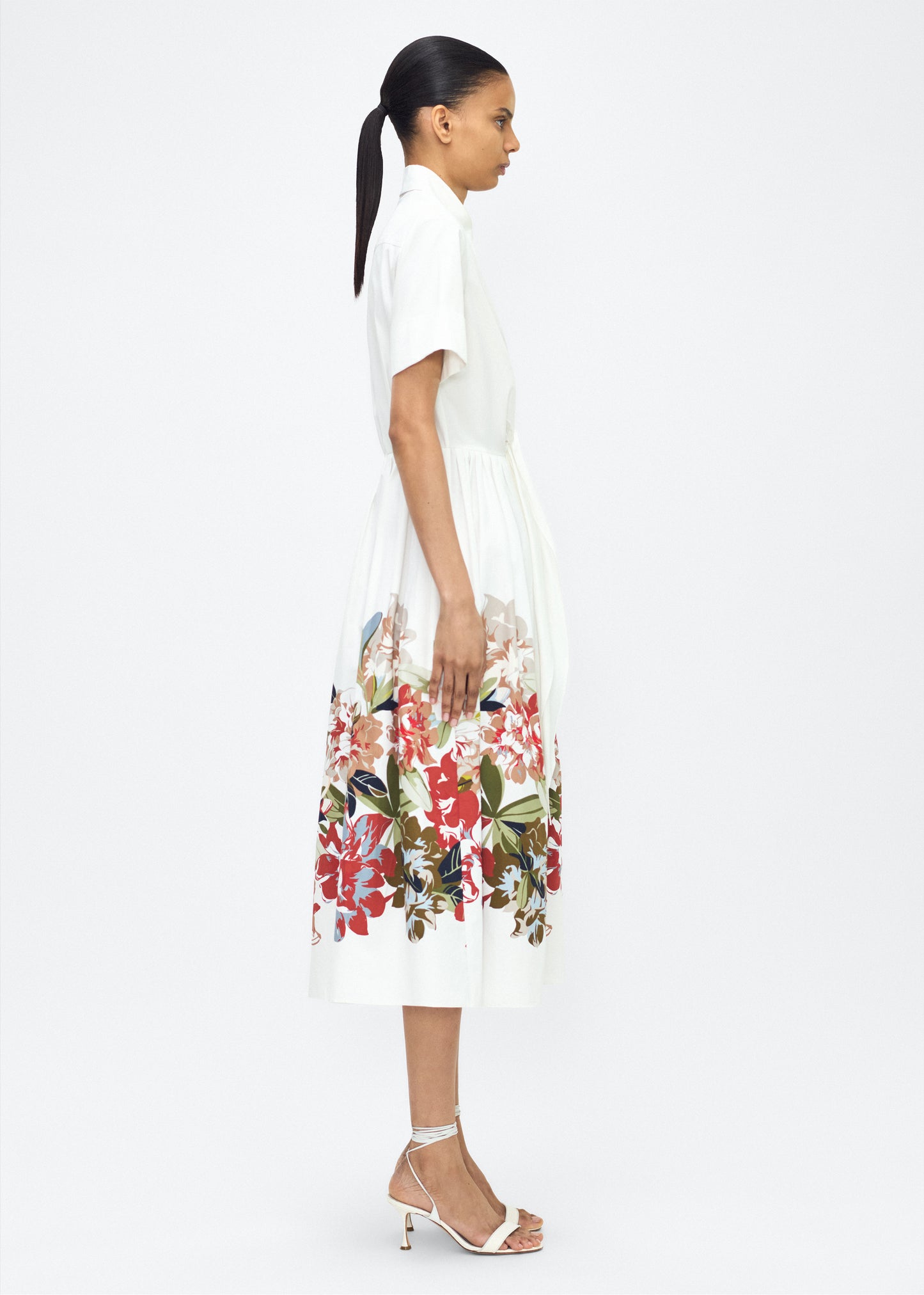 Model facing side Attacher Dress in white multi, P25 Collection from Adam Lippes; Our classic Attacher silhouette returns, crafted from Italian cotton poplin and printed with this season’s Sicily-inspired floral motif. Featuring side pockets and diamond monogram mother of pearl buttons, the attached waist sash can be tied at the front or back to cinch the waist.