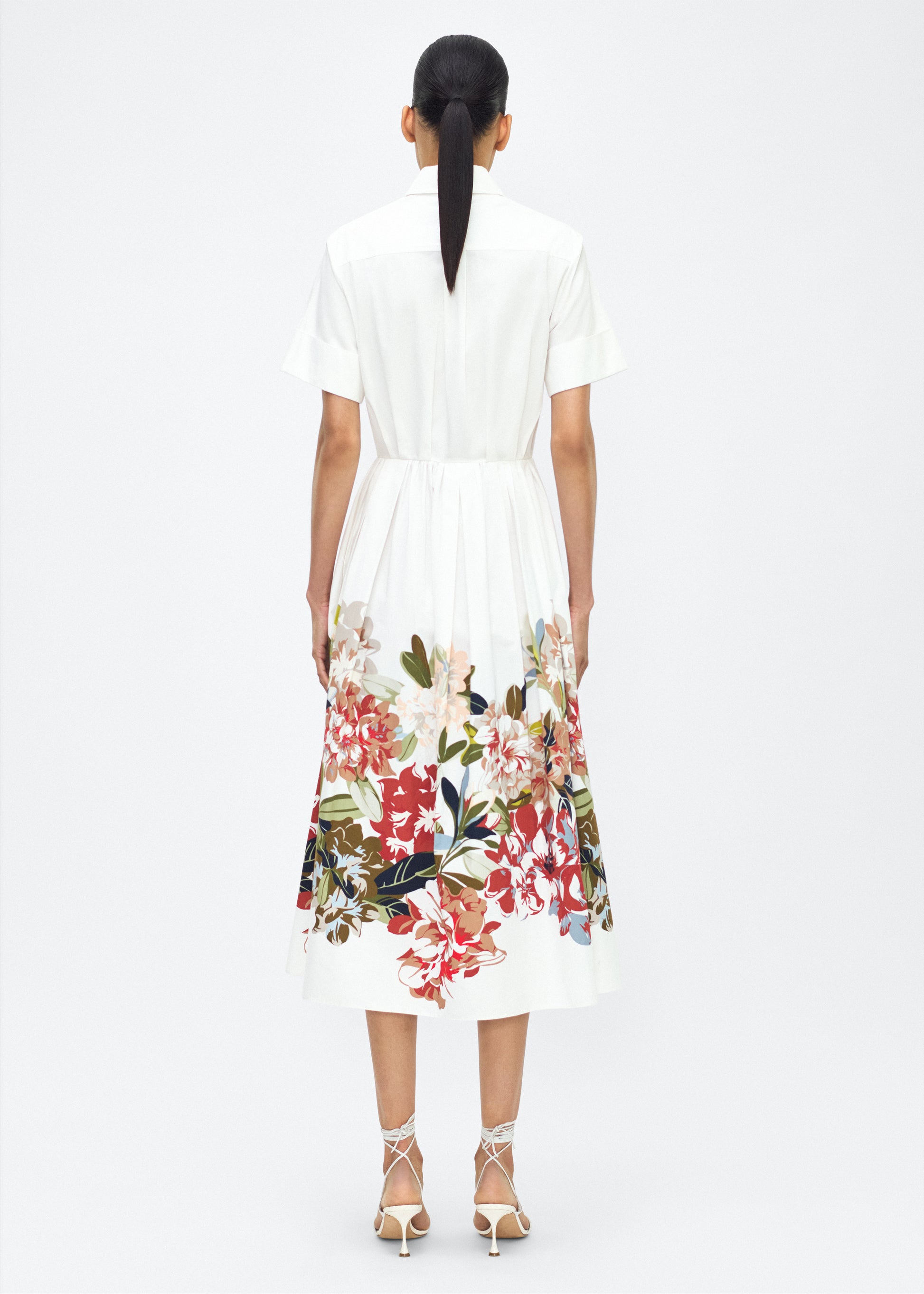Model facing back, Attacher Dress in white multi, P25 Collection from Adam Lippes; Our classic Attacher silhouette returns, crafted from Italian cotton poplin and printed with this season’s Sicily-inspired floral motif. Featuring side pockets and diamond monogram mother of pearl buttons, the attached waist sash can be tied at the front or back to cinch the waist.