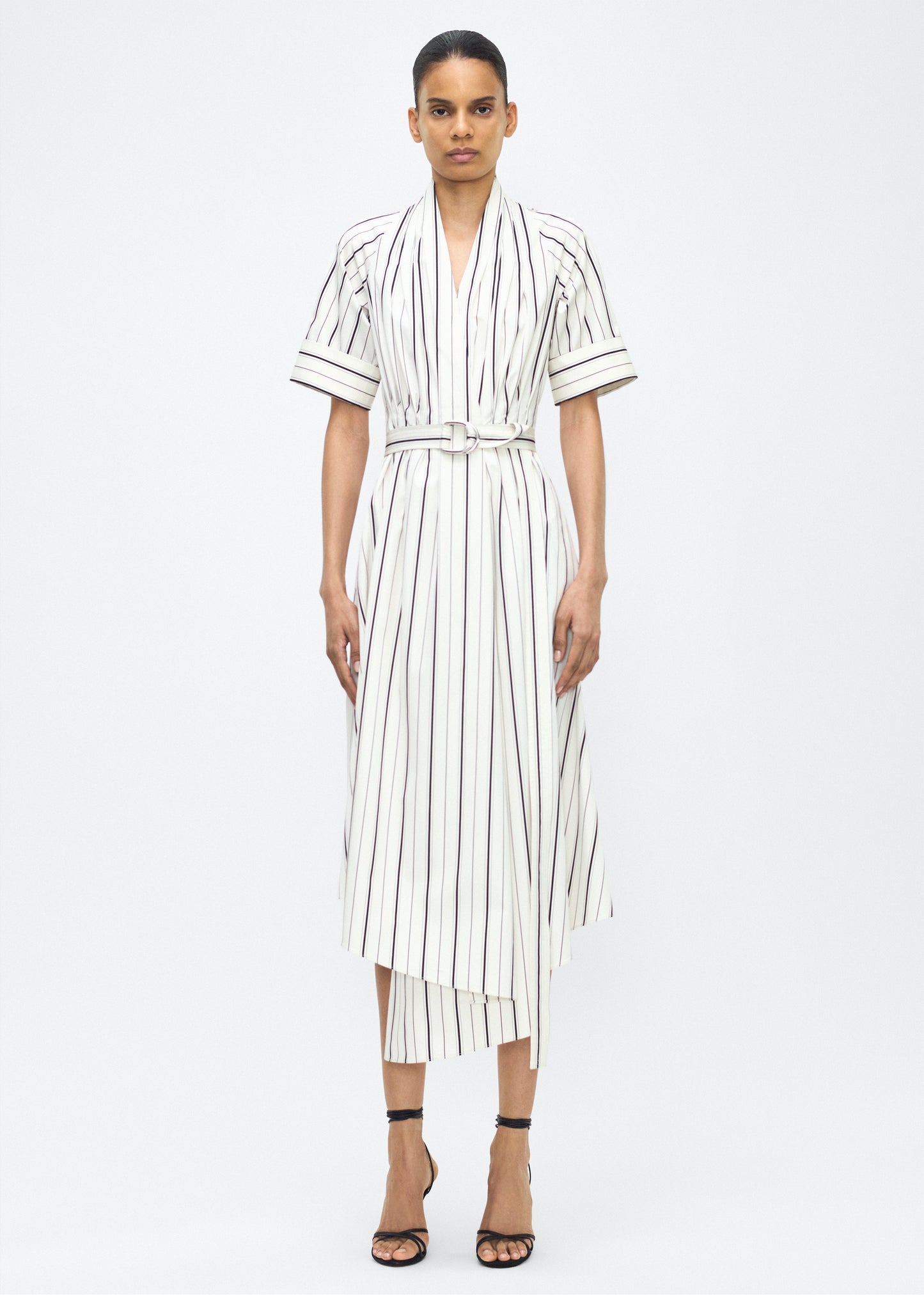 Model facing front, P25723SS, Ivory/Black, P25 Adam Lippes collection: The Nansi Dress is crafted from crisp stripe poplin in ivory and black, featuring an asymmetrical hem that adds a modern twist to a classic design. A sleek V-neckline and short sleeves enhance its effortless sophistication, while a fabric belt cinches the waist for a defined silhouette.