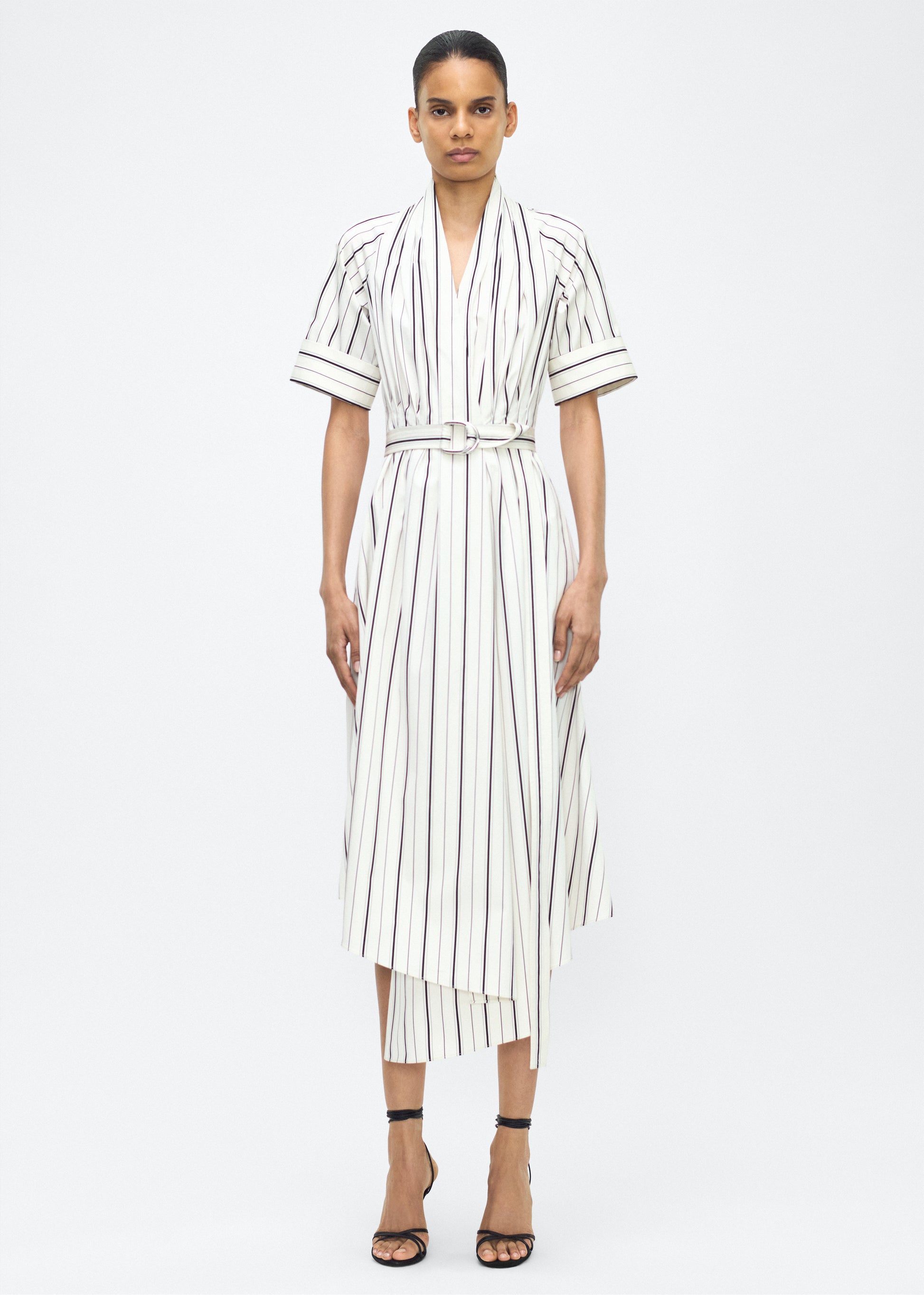 Model facing front, P25723SS, Ivory/Black, P25 Adam Lippes collection: The Nansi Dress is crafted from crisp stripe poplin in ivory and black, featuring an asymmetrical hem that adds a modern twist to a classic design. A sleek V-neckline and short sleeves enhance its effortless sophistication, while a fabric belt cinches the waist for a defined silhouette.