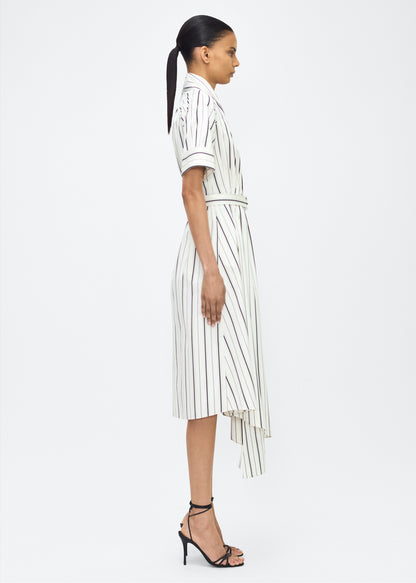 Model facing side, P25723SS, Ivory/Black, P25 Adam Lippes collection: The Nansi Dress is crafted from crisp stripe poplin in ivory and black, featuring an asymmetrical hem that adds a modern twist to a classic design. A sleek V-neckline and short sleeves enhance its effortless sophistication, while a fabric belt cinches the waist for a defined silhouette.