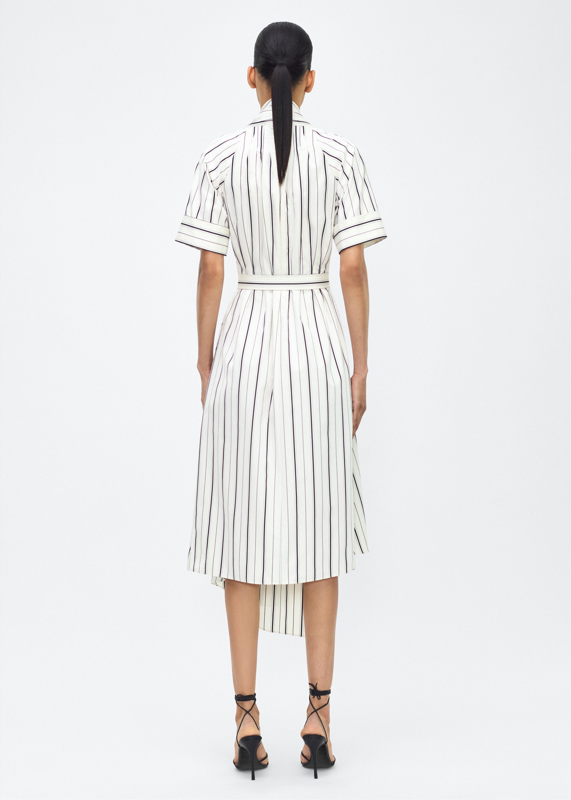Model facing back, P25723SS, Ivory/Black, P25 Adam Lippes collection: The Nansi Dress is crafted from crisp stripe poplin in ivory and black, featuring an asymmetrical hem that adds a modern twist to a classic design. A sleek V-neckline and short sleeves enhance its effortless sophistication, while a fabric belt cinches the waist for a defined silhouette.