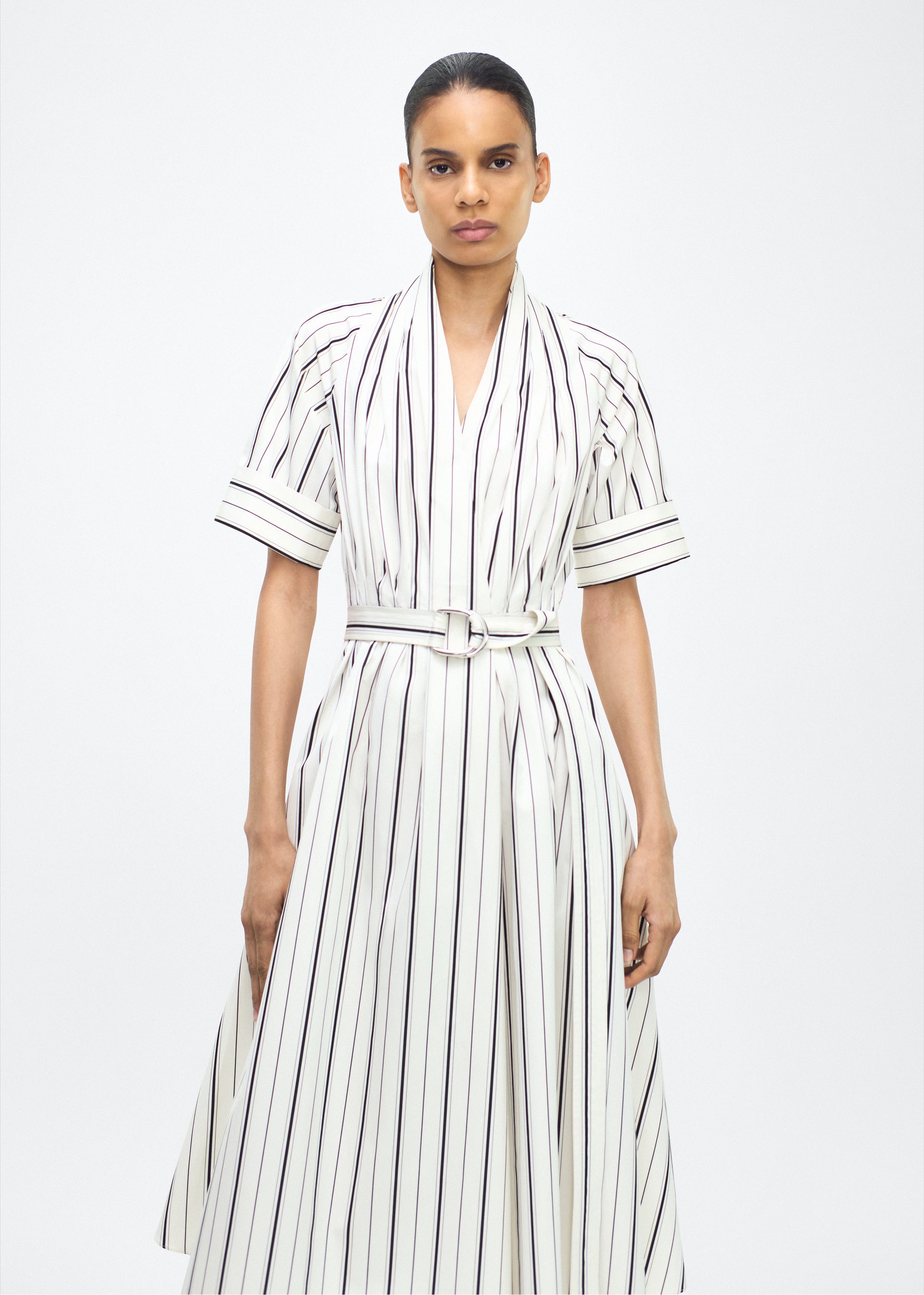 Model facing front, P25723SS, Ivory/Black, P25 Adam Lippes collection: The Nansi Dress is crafted from crisp stripe poplin in ivory and black, featuring an asymmetrical hem that adds a modern twist to a classic design. A sleek V-neckline and short sleeves enhance its effortless sophistication, while a fabric belt cinches the waist for a defined silhouette.