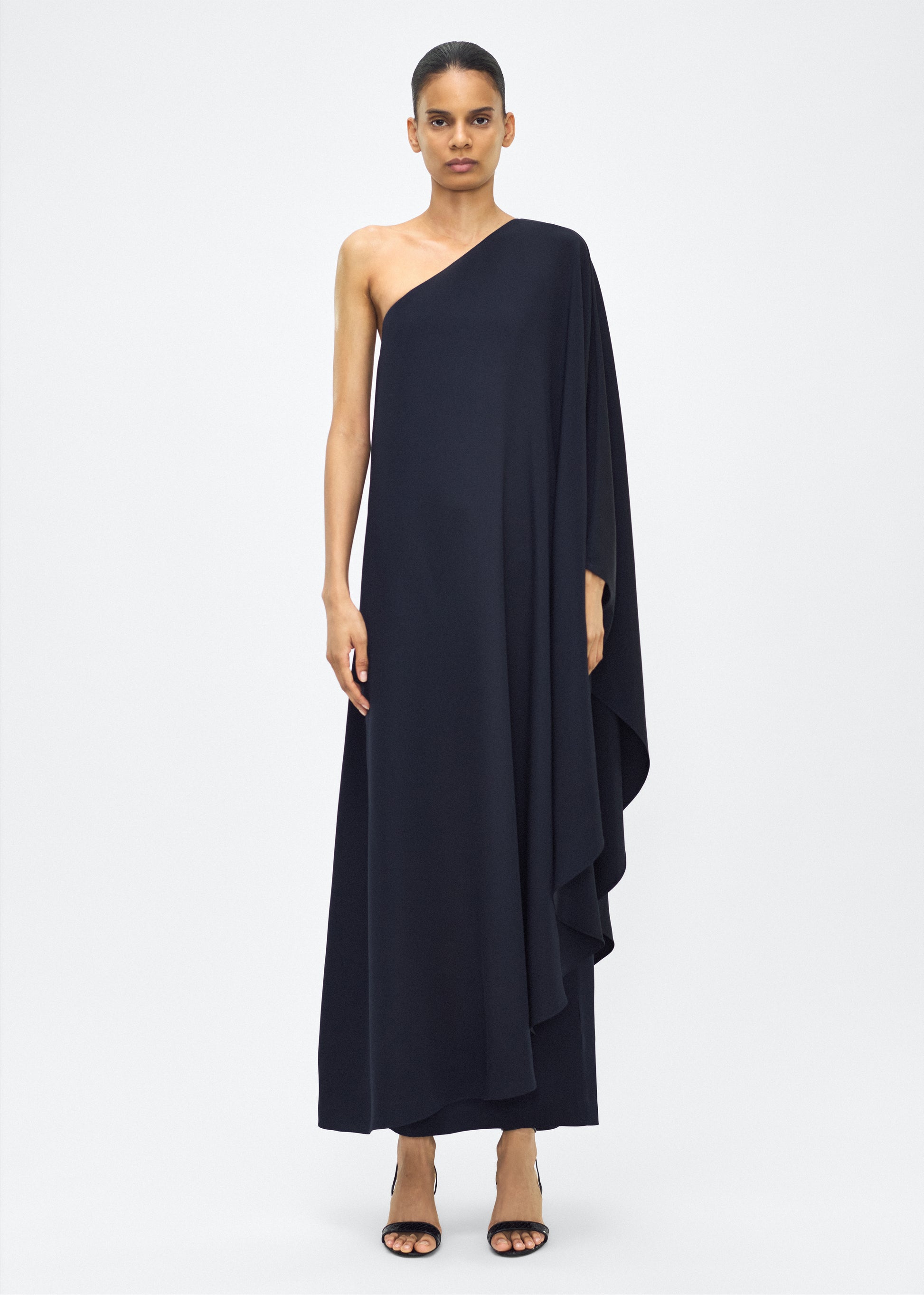 Image of Model facing front, P25747SE, Navy, Adam Lippes P25 Collection; The Solene Dress is a striking one-shoulder design crafted from luxurious silk crepe. The fabric drapes gracefully, creating an elegant, fluid silhouette. The asymmetrical cut is complemented by the soft, flowing lines that cascade effortlessly.