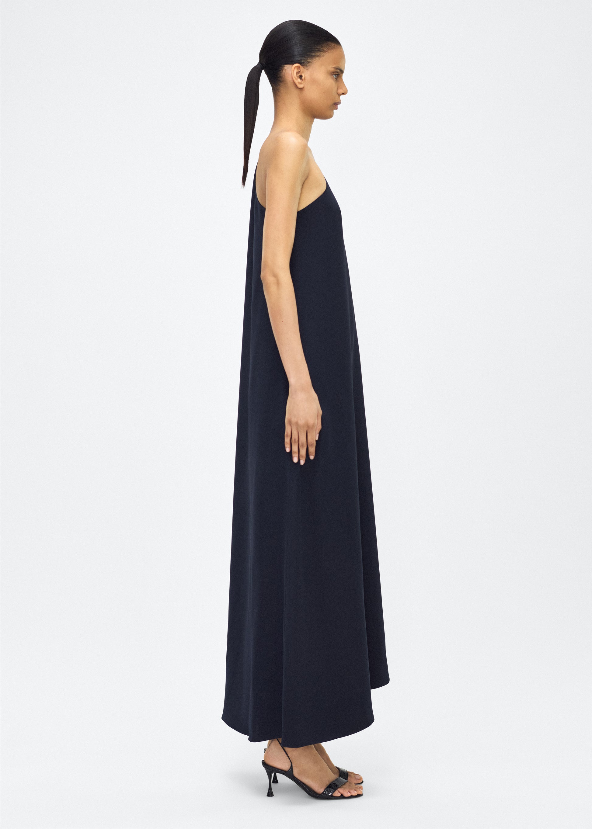 Image of Model facing side, P25747SE, Navy, Adam Lippes P25 Collection; The Solene Dress is a striking one-shoulder design crafted from luxurious silk crepe. The fabric drapes gracefully, creating an elegant, fluid silhouette. The asymmetrical cut is complemented by the soft, flowing lines that cascade effortlessly.