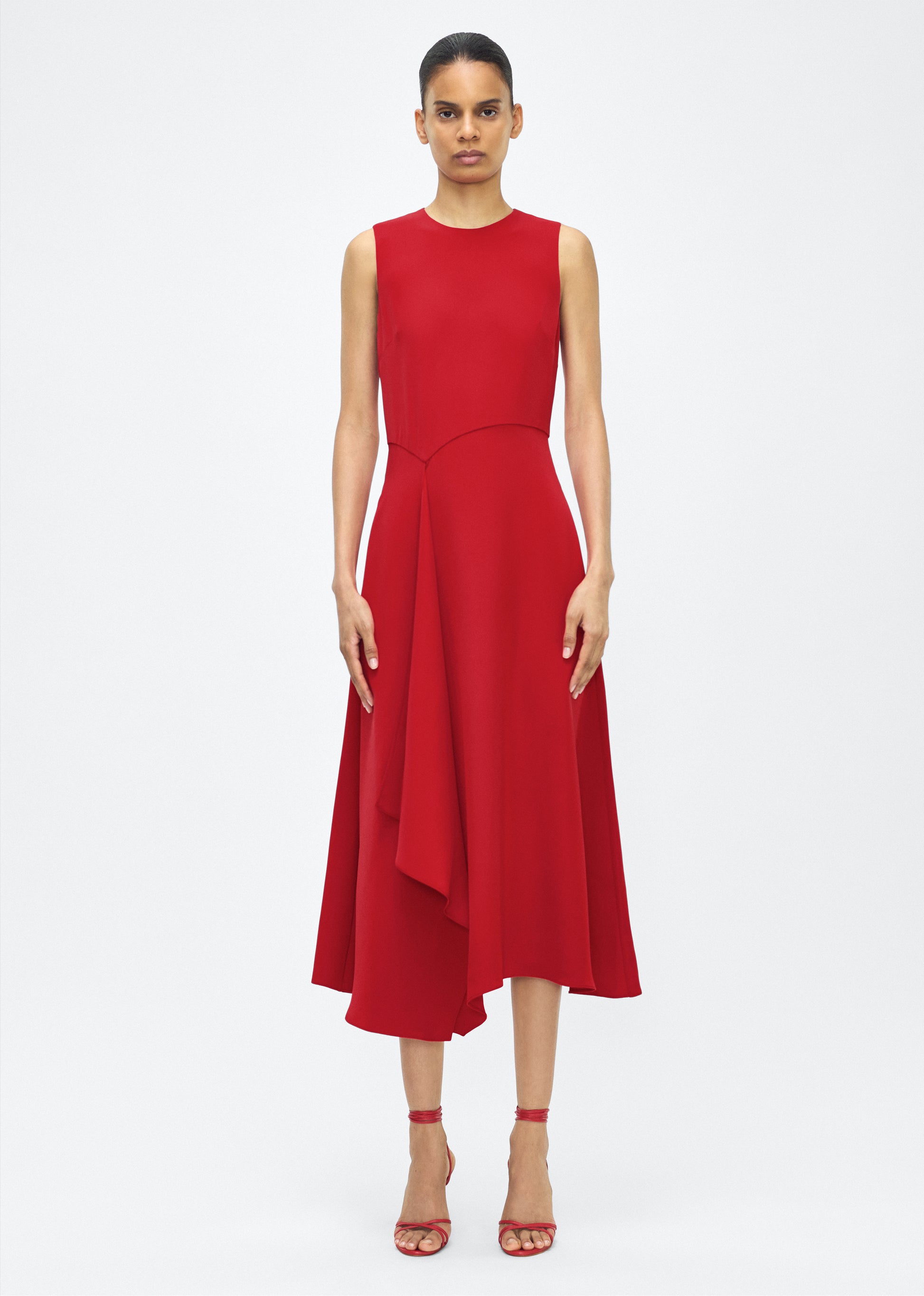 Model facing front– Lynn Dress, P25753SE, Poppy – Adam Lippes, P25 collection: The Lynn Dress introduces a striking new silhouette in silk crepe. This sleeveless design features a softly contoured waistline and an asymmetric draped panel, creating a dynamic and effortless shape.