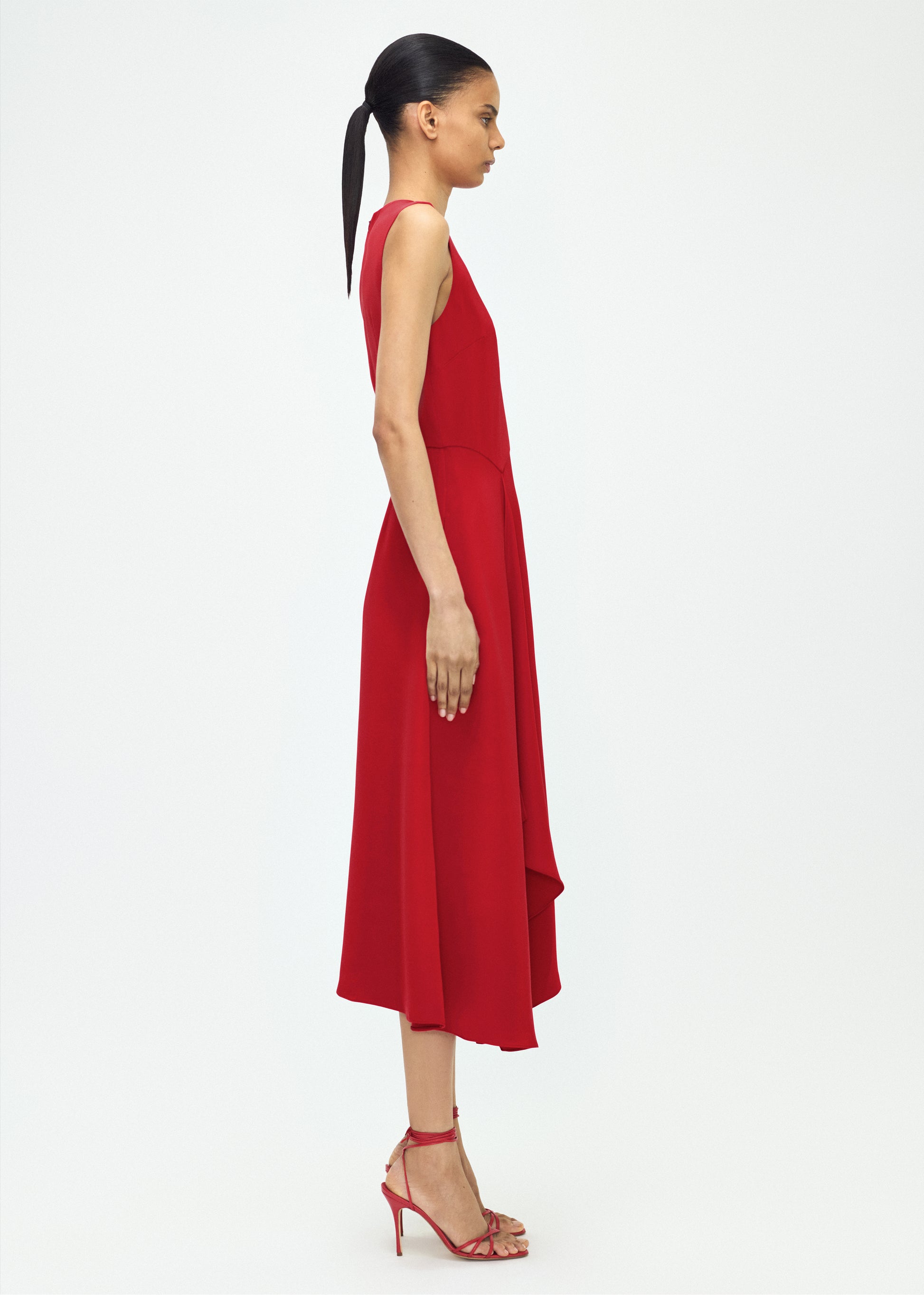 Model facing side- Lynn Dress, P25753SE, Poppy – Adam Lippes, P25 collection: The Lynn Dress introduces a striking new silhouette in silk crepe. This sleeveless design features a softly contoured waistline and an asymmetric draped panel, creating a dynamic and effortless shape.