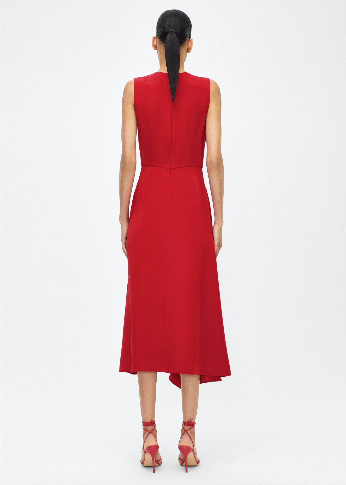 Model facing back, Lynn Dress, P25753SE, Poppy – Adam Lippes, P25 collection: The Lynn Dress introduces a striking new silhouette in silk crepe. This sleeveless design features a softly contoured waistline and an asymmetric draped panel, creating a dynamic and effortless shape.