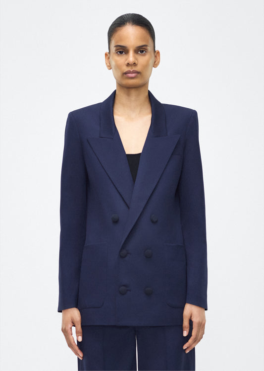 Model image front – Riva jacket, P25805TW, navy – Adam Lippes, P25 collection: The Riva Jacket debuts in lightweight tropical wool—a structured, resilient fabric that maintains its shape while offering ease of movement. Featuring peaked lapels and a double-breasted front, it’s finished with covered buttons for a refined, polished look.
