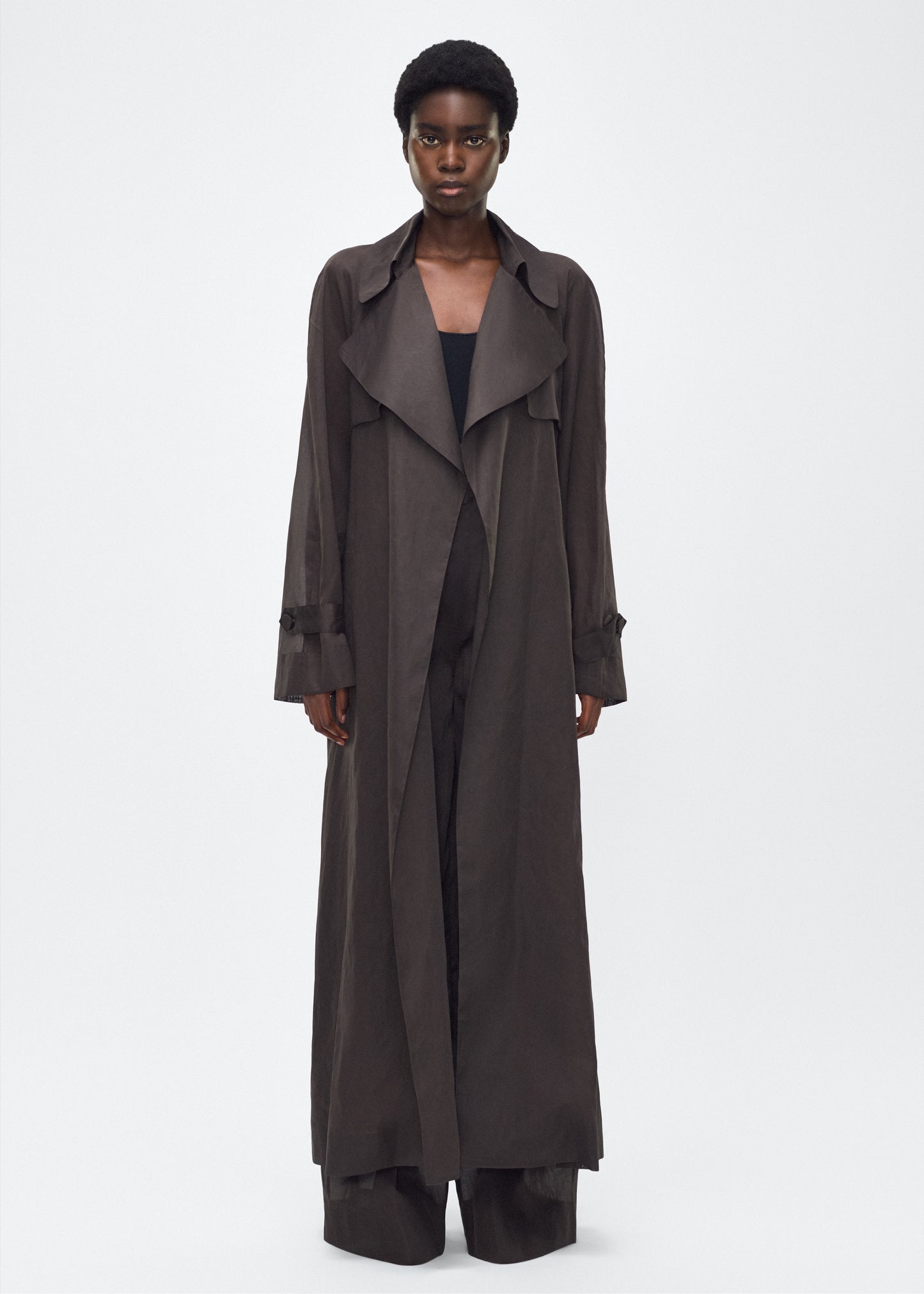 Image of model facing front, wearing the Jupiter Trench, P25 Adam Lippes Collection, color Ebony. The Jupiter Trench is crafted from lightweight cotton organdy, offering a semi-sheer, airy feel. Finished with the Adam Lippes diamond motif, embroidered in silk charmeuse at the back, and designed with a belt to cinch the waist, creating a sleek silhouette with considered details.