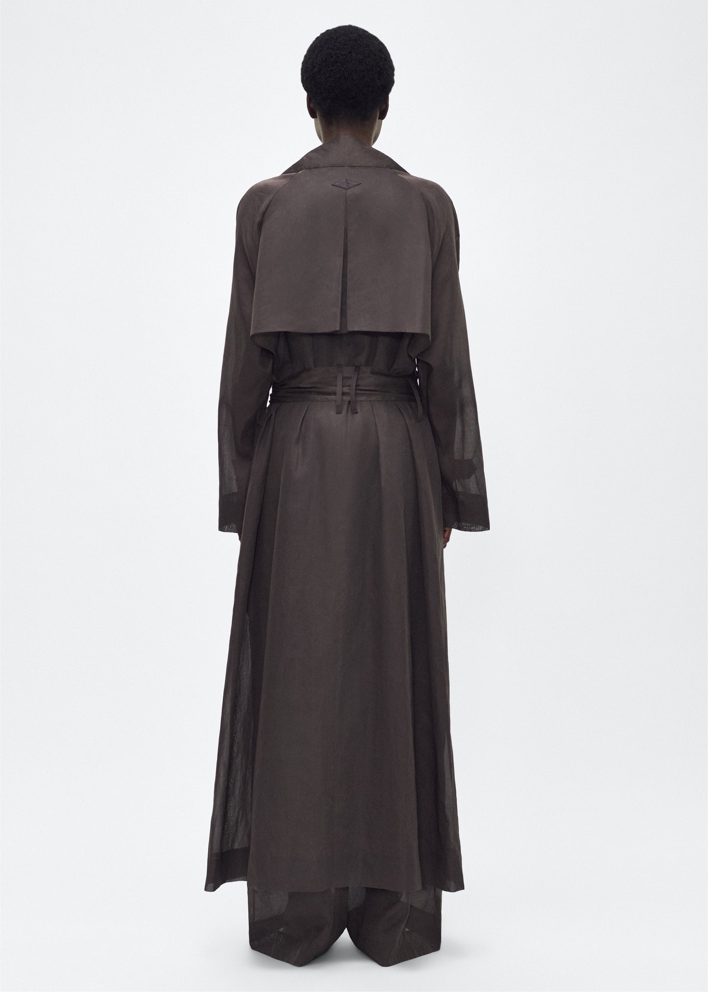 Image of model facing back, wearing the Jupiter Trench, P25 Adam Lippes Collection, color Ebony. The Jupiter Trench is crafted from lightweight cotton organdy, offering a semi-sheer, airy feel. Finished with the Adam Lippes diamond motif, embroidered in silk charmeuse at the back, and designed with a belt to cinch the waist, creating a sleek silhouette with considered details.