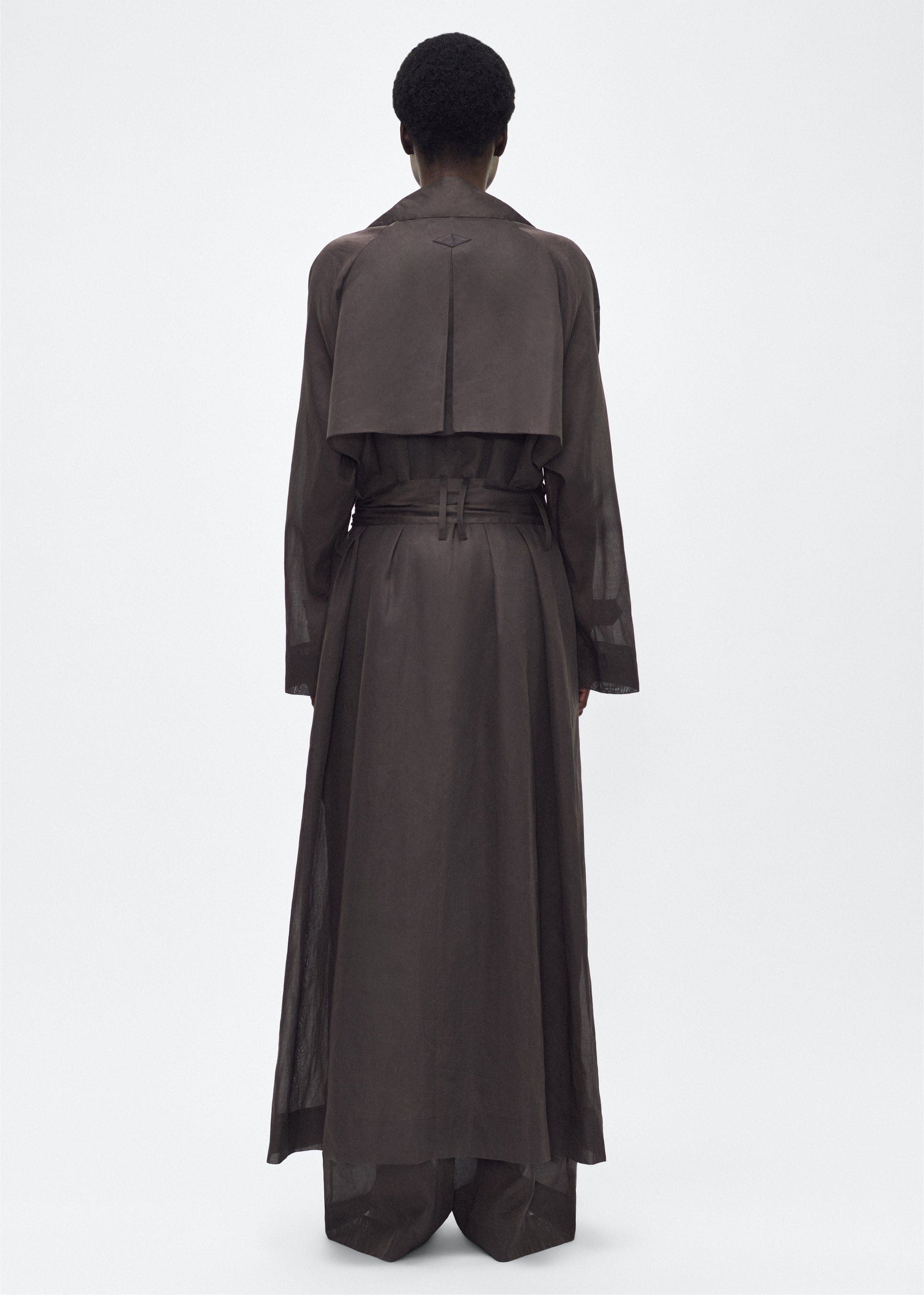 Image of model facing back, wearing the Jupiter Trench, P25 Adam Lippes Collection, color Ebony. The Jupiter Trench is crafted from lightweight cotton organdy, offering a semi-sheer, airy feel. Finished with the Adam Lippes diamond motif, embroidered in silk charmeuse at the back, and designed with a belt to cinch the waist, creating a sleek silhouette with considered details.