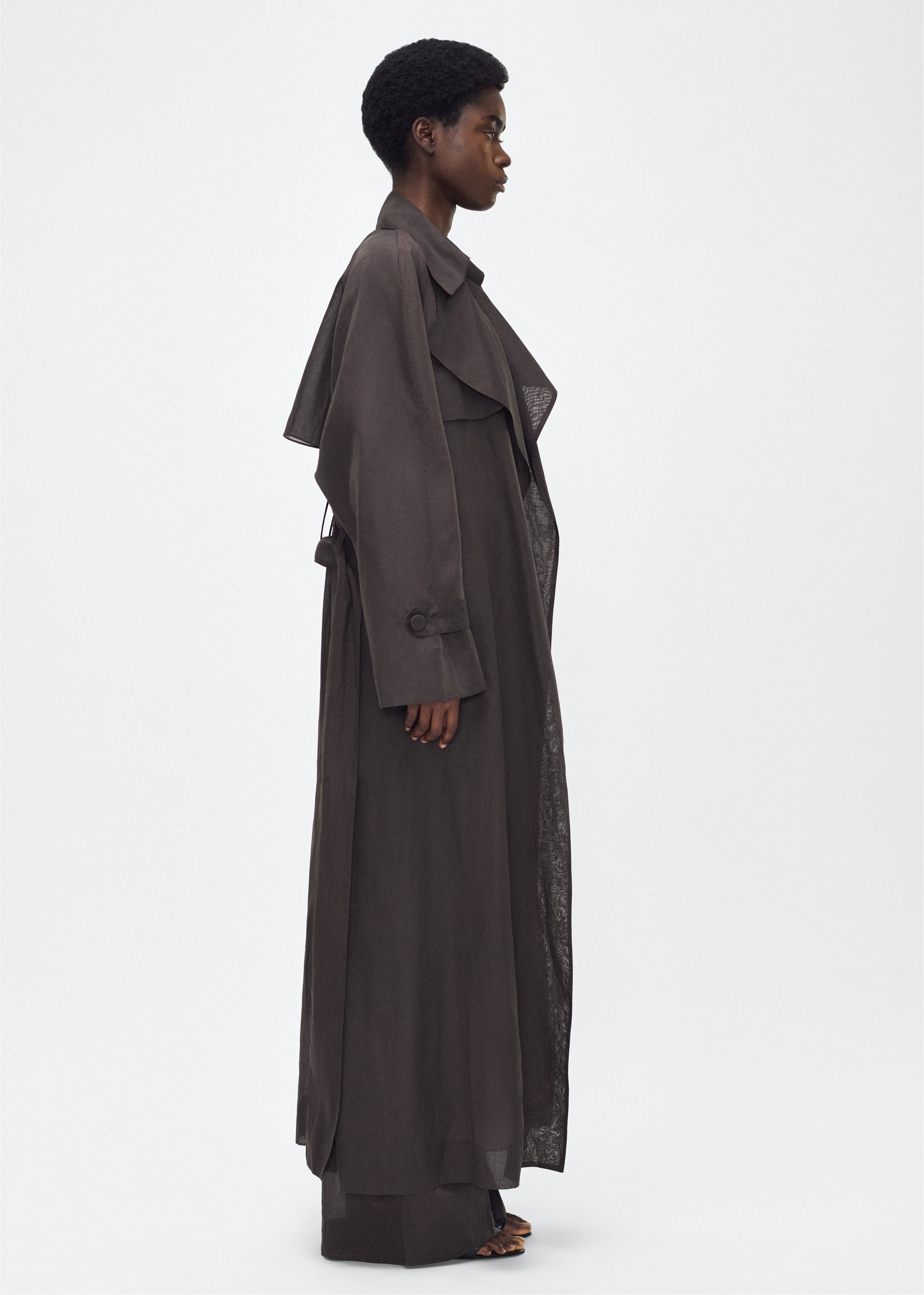 Image of model facing side, wearing the Jupiter Trench, P25 Adam Lippes Collection, color Ebony. The Jupiter Trench is crafted from lightweight cotton organdy, offering a semi-sheer, airy feel. Finished with the Adam Lippes diamond motif, embroidered in silk charmeuse at the back, and designed with a belt to cinch the waist, creating a sleek silhouette with considered details.