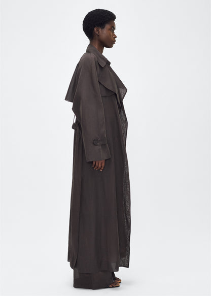 Image of model facing side, wearing the Jupiter Trench, P25 Adam Lippes Collection, color Ebony. The Jupiter Trench is crafted from lightweight cotton organdy, offering a semi-sheer, airy feel. Finished with the Adam Lippes diamond motif, embroidered in silk charmeuse at the back, and designed with a belt to cinch the waist, creating a sleek silhouette with considered details.