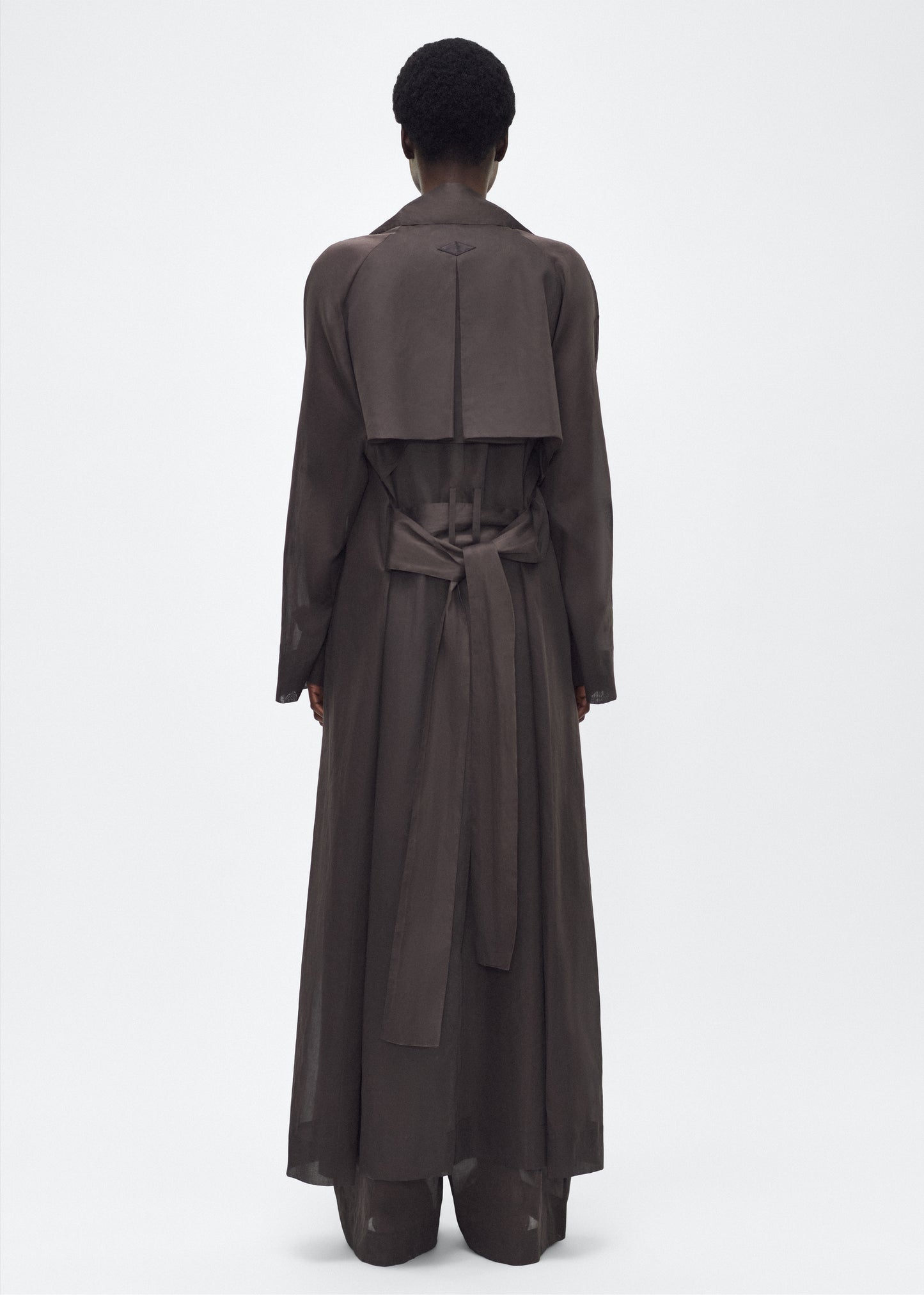 Image of model facing back, wearing the Jupiter Trench, P25 Adam Lippes Collection, color Ebony. The Jupiter Trench is crafted from lightweight cotton organdy, offering a semi-sheer, airy feel. Finished with the Adam Lippes diamond motif, embroidered in silk charmeuse at the back, and designed with a belt to cinch the waist, creating a sleek silhouette with considered details.