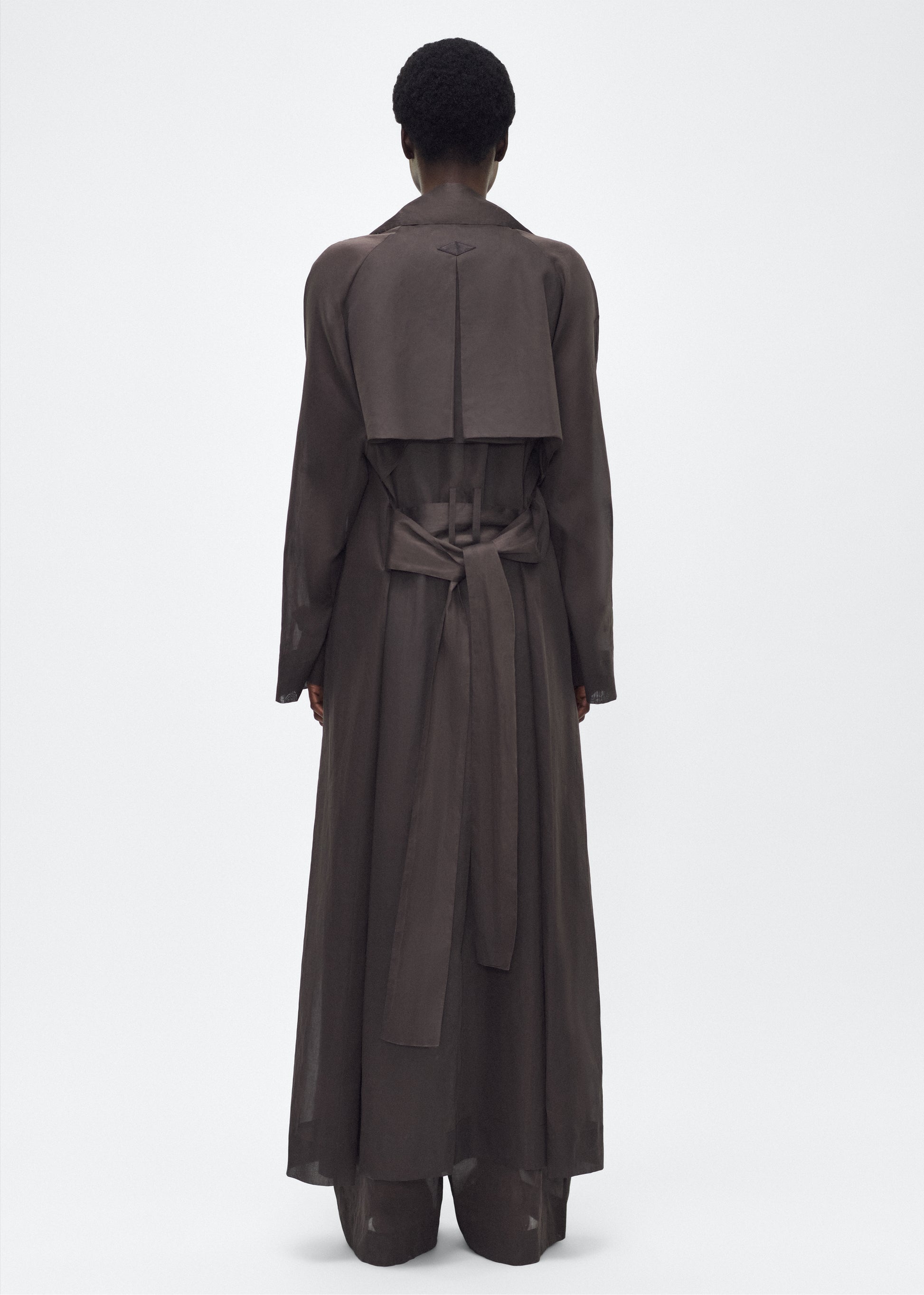 Image of model facing back, wearing the Jupiter Trench, P25 Adam Lippes Collection, color Ebony. The Jupiter Trench is crafted from lightweight cotton organdy, offering a semi-sheer, airy feel. Finished with the Adam Lippes diamond motif, embroidered in silk charmeuse at the back, and designed with a belt to cinch the waist, creating a sleek silhouette with considered details.