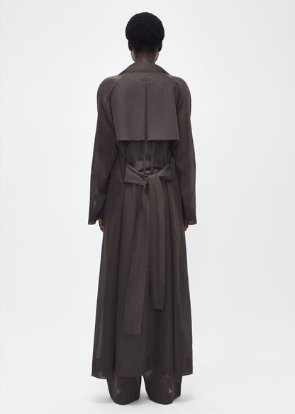 Image of model facing back, wearing the Jupiter Trench, P25 Adam Lippes Collection, color Ebony. The Jupiter Trench is crafted from lightweight cotton organdy, offering a semi-sheer, airy feel. Finished with the Adam Lippes diamond motif, embroidered in silk charmeuse at the back, and designed with a belt to cinch the waist, creating a sleek silhouette with considered details.