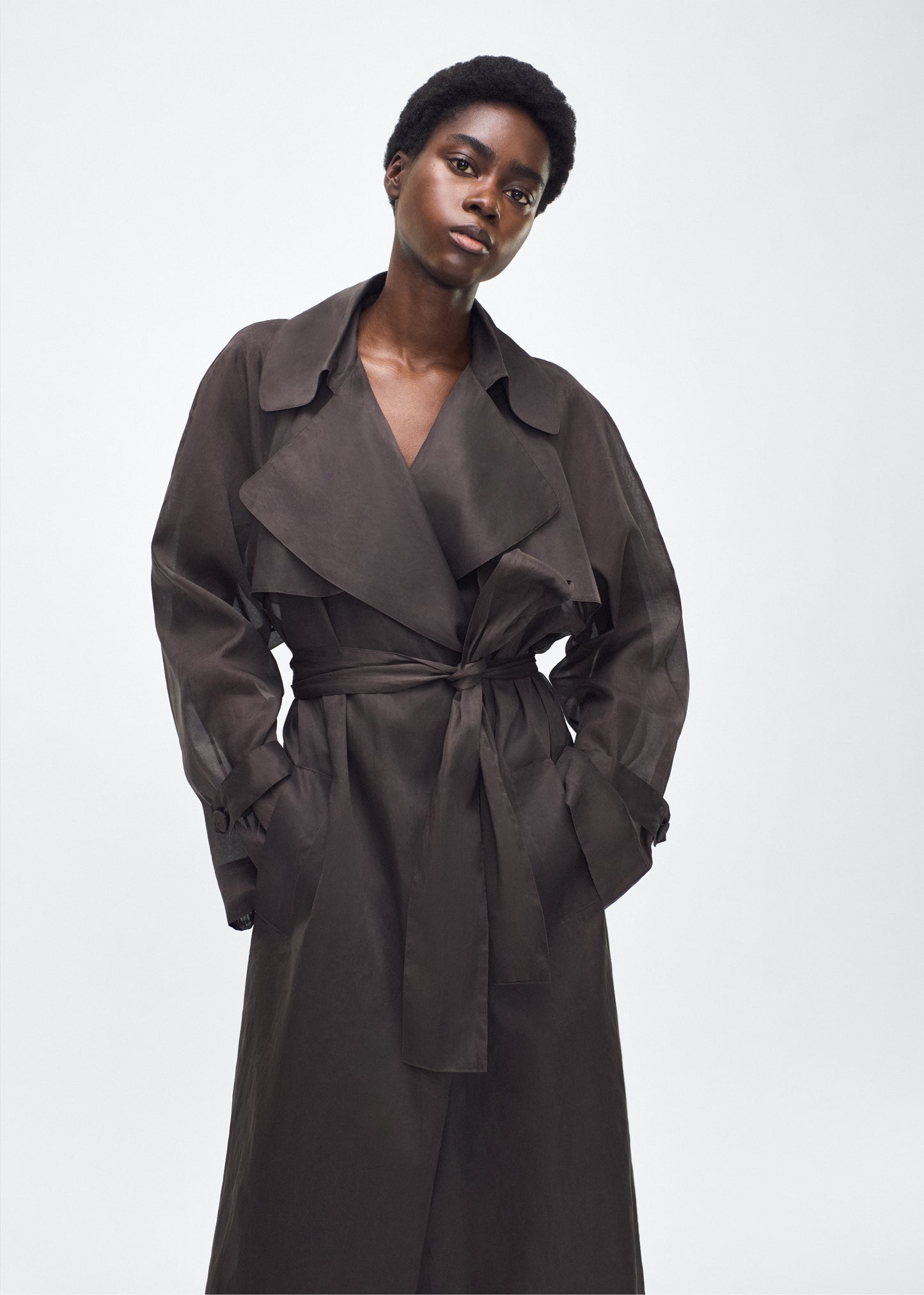 Image of model facing front, wearing the Jupiter Trench, P25 Adam Lippes Collection, color Ebony. The Jupiter Trench is crafted from lightweight cotton organdy, offering a semi-sheer, airy feel. Finished with the Adam Lippes diamond motif, embroidered in silk charmeuse at the back, and designed with a belt to cinch the waist, creating a sleek silhouette with considered details.
