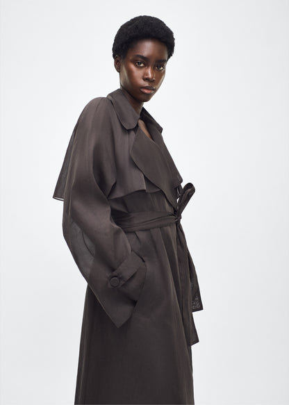 Image of model facing side, wearing the Jupiter Trench, P25 Adam Lippes Collection, color Ebony. The Jupiter Trench is crafted from lightweight cotton organdy, offering a semi-sheer, airy feel. Finished with the Adam Lippes diamond motif, embroidered in silk charmeuse at the back, and designed with a belt to cinch the waist, creating a sleek silhouette with considered details.