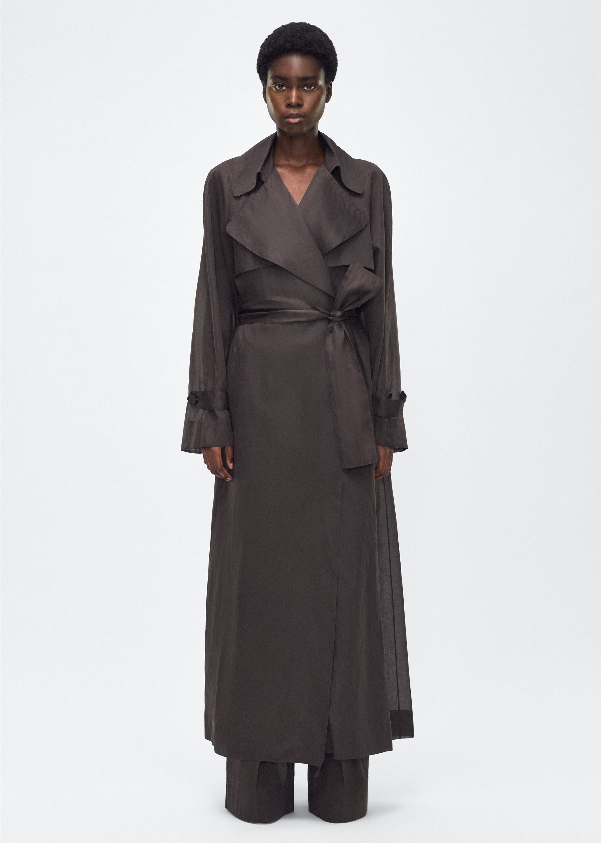Image of model facing front, wearing the Jupiter Trench, P25 Adam Lippes Collection, color Ebony. The Jupiter Trench is crafted from lightweight cotton organdy, offering a semi-sheer, airy feel. Finished with the Adam Lippes diamond motif, embroidered in silk charmeuse at the back, and designed with a belt to cinch the waist, creating a sleek silhouette with considered details.