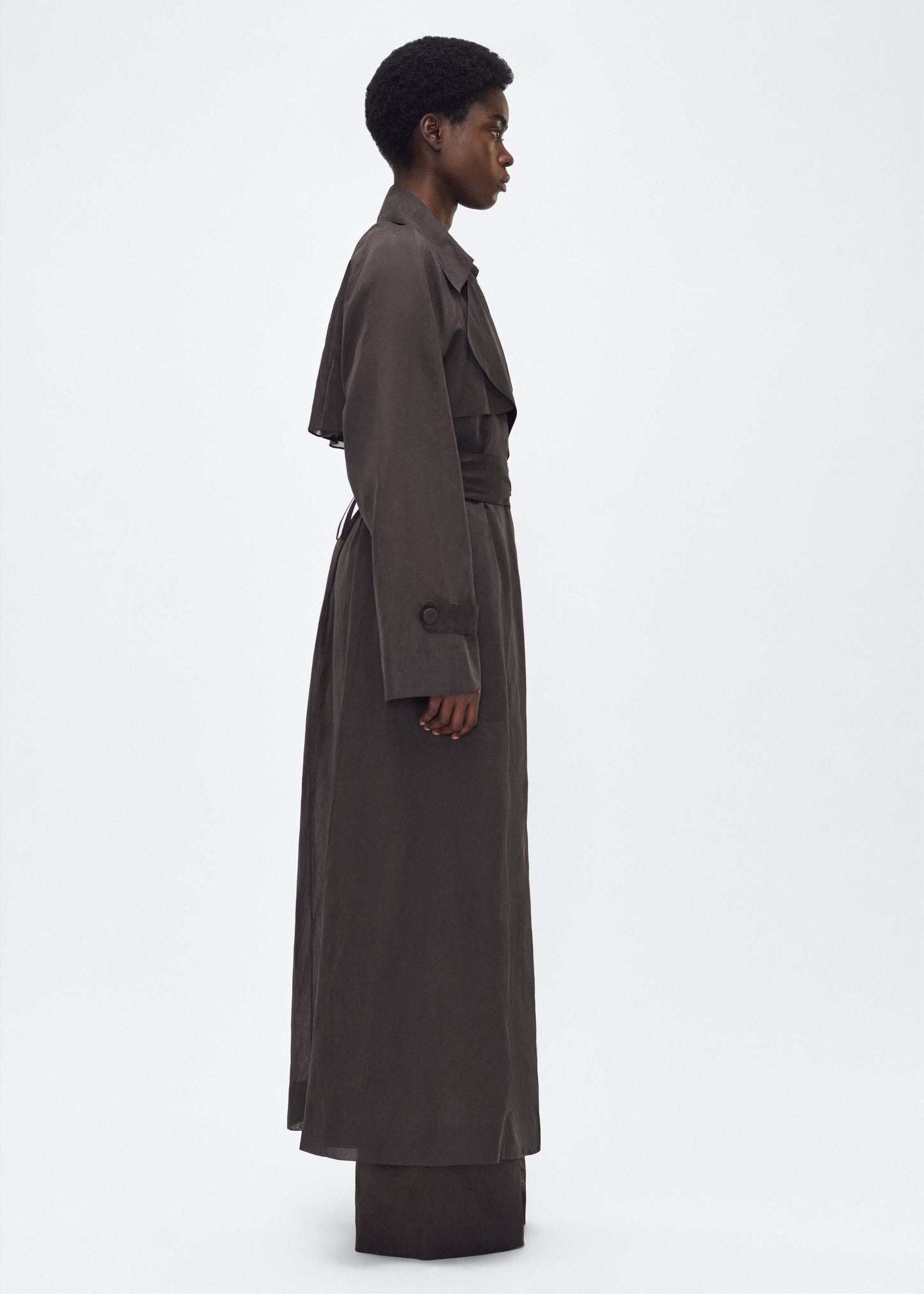 Image of model facing side, wearing the Jupiter Trench, P25 Adam Lippes Collection, color Ebony. The Jupiter Trench is crafted from lightweight cotton organdy, offering a semi-sheer, airy feel. Finished with the Adam Lippes diamond motif, embroidered in silk charmeuse at the back, and designed with a belt to cinch the waist, creating a sleek silhouette with considered details.