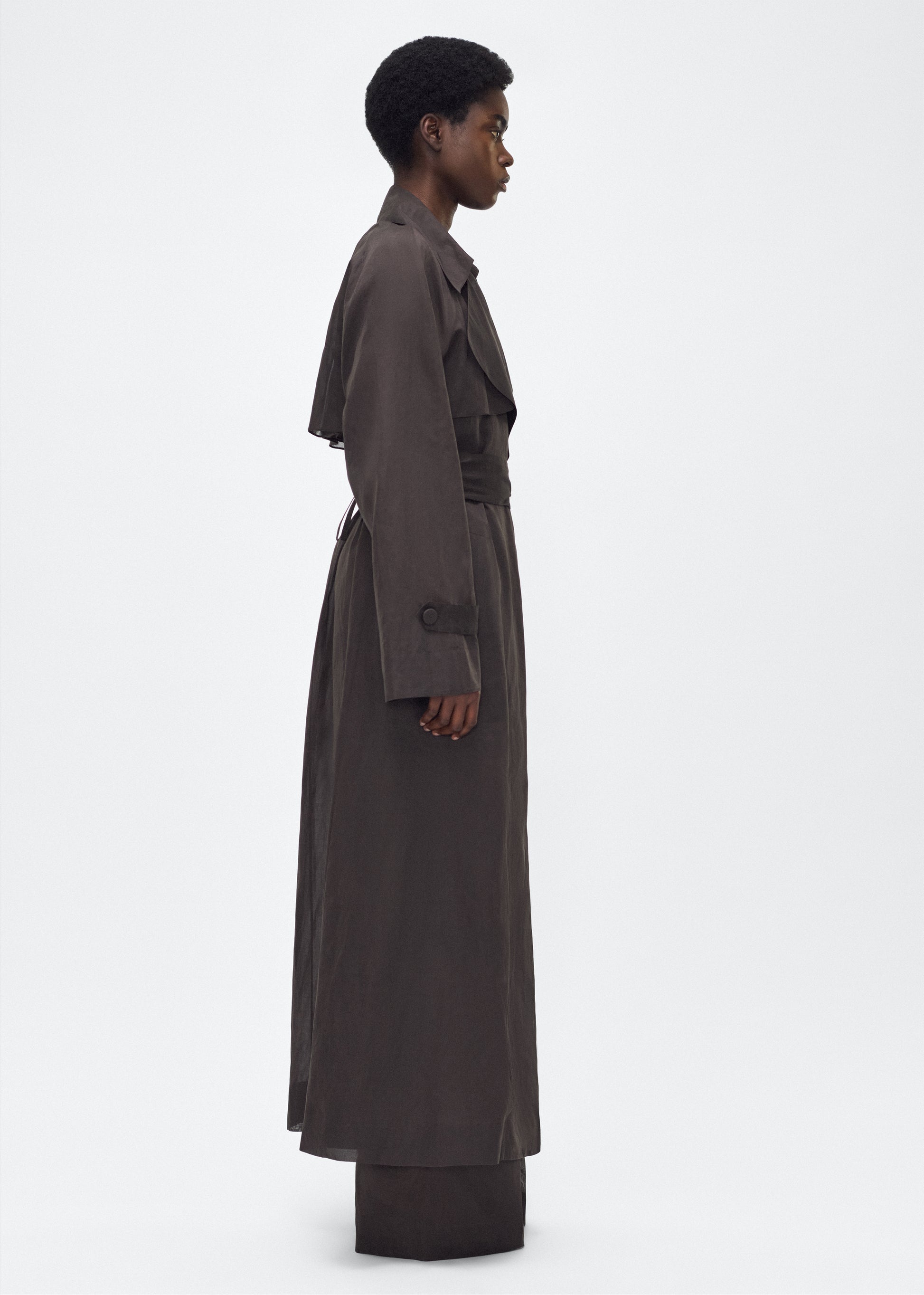 Image of model facing side, wearing the Jupiter Trench, P25 Adam Lippes Collection, color Ebony. The Jupiter Trench is crafted from lightweight cotton organdy, offering a semi-sheer, airy feel. Finished with the Adam Lippes diamond motif, embroidered in silk charmeuse at the back, and designed with a belt to cinch the waist, creating a sleek silhouette with considered details.