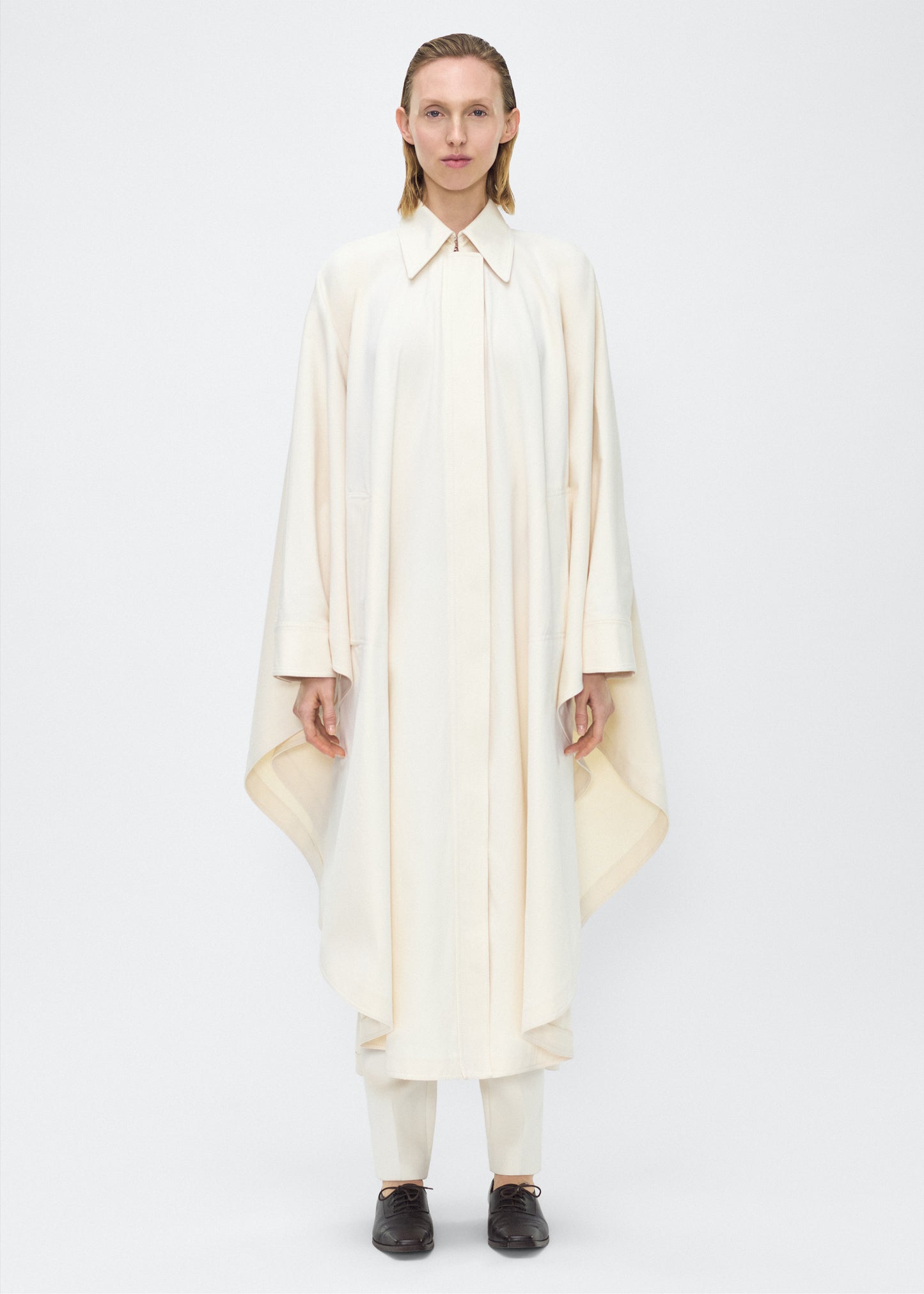 Image of Model facing front, wearing the Langston Poncho, P25914CT, in color ivory, Adam Lippes P25 collection; The Langston Poncho in ivory cotton twill offers an elegant take on the classic poncho. With its modern, relaxed shape, sharp tailoring, and hidden corozo button closure down the center front, it effortlessly elevates any ensemble. The ivory hue and clean lines are complemented by thoughtful details.