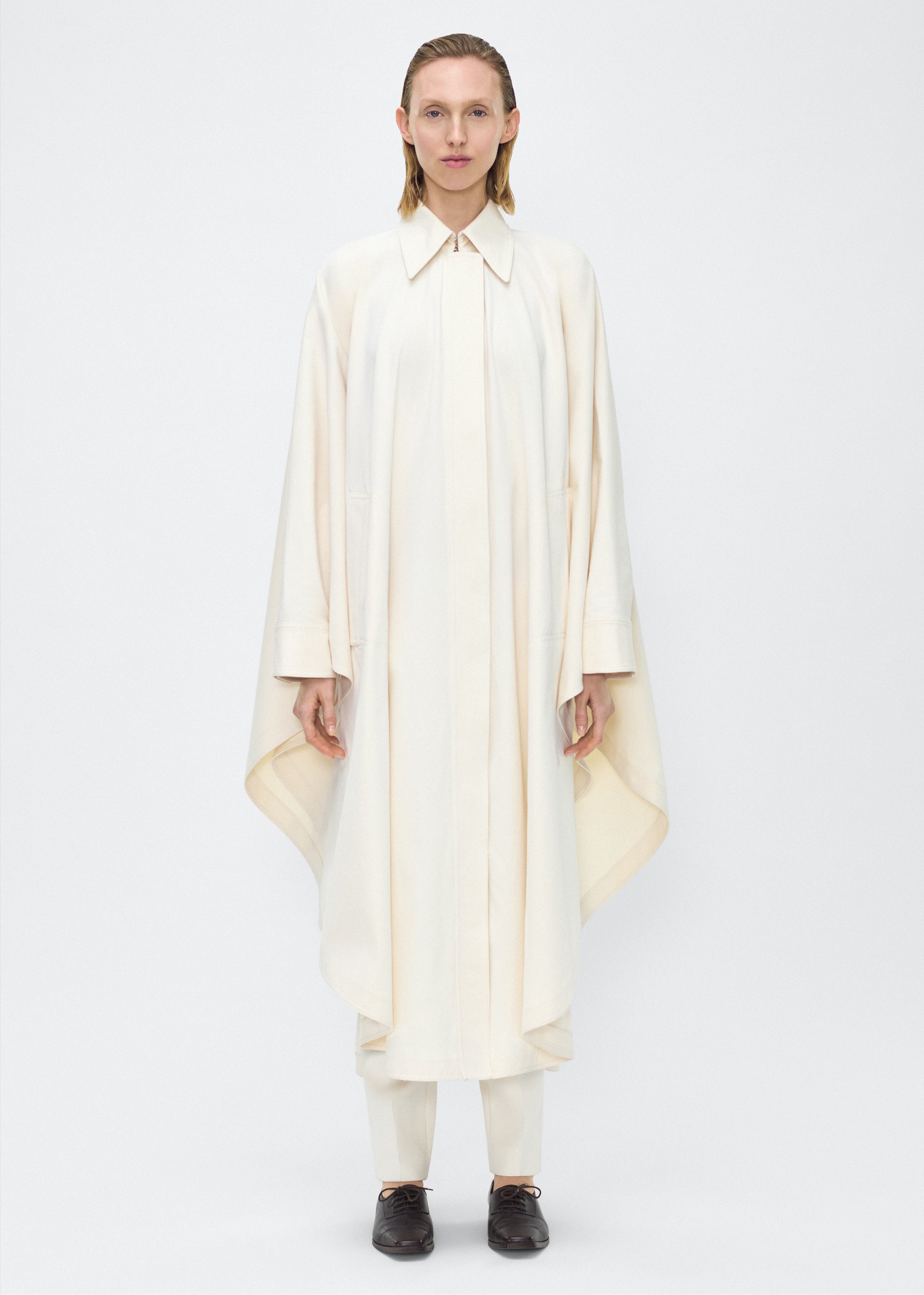Image of Model facing front, wearing the Langston Poncho, P25914CT, in color ivory, Adam Lippes P25 collection; The Langston Poncho in ivory cotton twill offers an elegant take on the classic poncho. With its modern, relaxed shape, sharp tailoring, and hidden corozo button closure down the center front, it effortlessly elevates any ensemble. The ivory hue and clean lines are complemented by thoughtful details.
