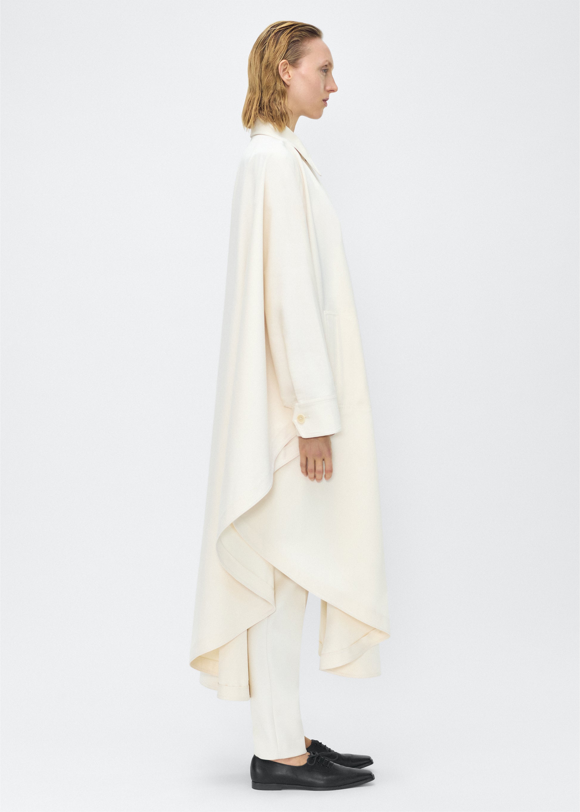 Image of Model facing side, wearing the Langston Poncho, P25914CT, in color ivory, Adam Lippes P25 collection; The Langston Poncho in ivory cotton twill offers an elegant take on the classic poncho. With its modern, relaxed shape, sharp tailoring, and hidden corozo button closure down the center front, it effortlessly elevates any ensemble. The ivory hue and clean lines are complemented by thoughtful details.