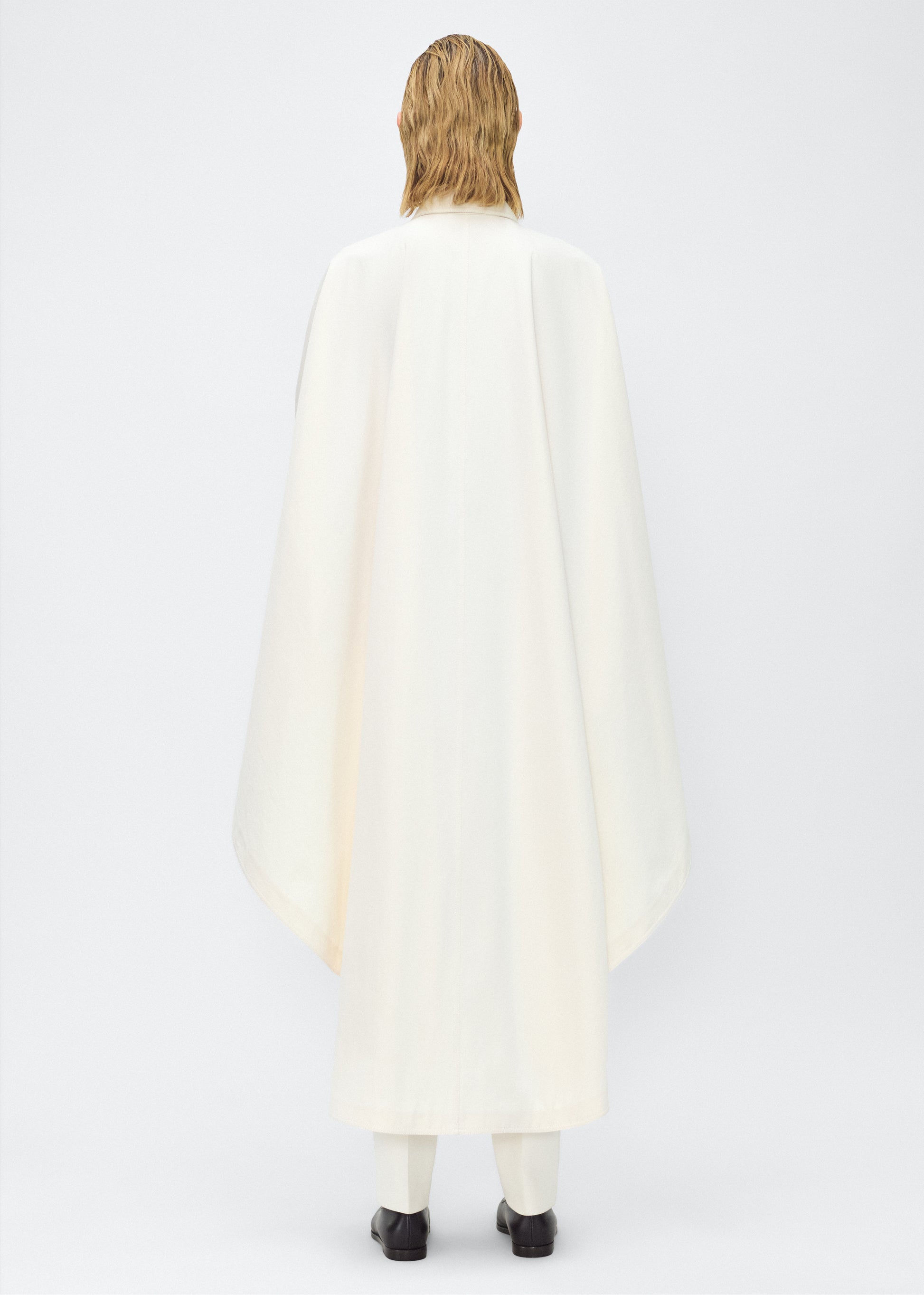 Image of Model facing back, wearing the Langston Poncho, P25914CT, in color ivory, Adam Lippes P25 collection; The Langston Poncho in ivory cotton twill offers an elegant take on the classic poncho. With its modern, relaxed shape, sharp tailoring, and hidden corozo button closure down the center front, it effortlessly elevates any ensemble. The ivory hue and clean lines are complemented by thoughtful details.