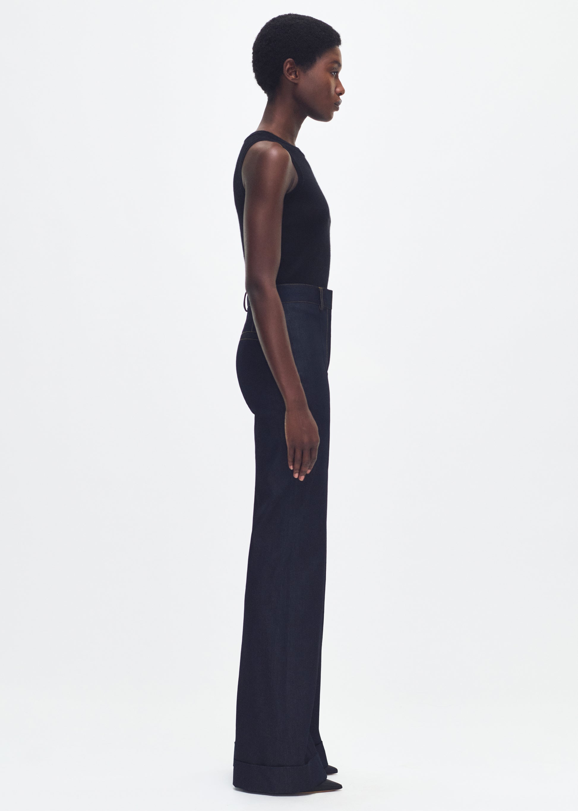 Side facing image of a model wearing a black tank top tucked into dark denim jeans. 